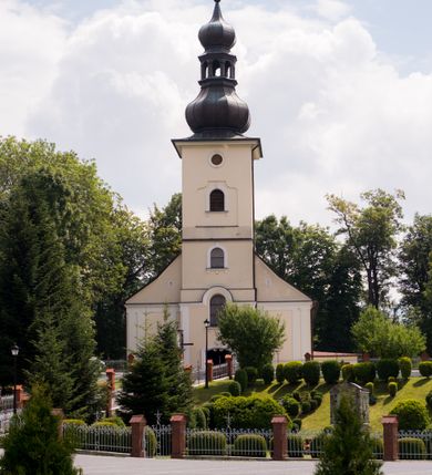 Zdjęcie nr 12: Kościół, zwrócony w kierunku południowo-wschodnim, jest budowlą murowaną z cegły i kamienia, na rzucie podłużnym. Długość bryły wynosi 35, a szerokość 16 metrów. Korpus nawowy jest trójnawowy i czteroprzęsłowy. Stosunkowo szerokie, jednoprzęsłowe, zamknięte półkoliście prezbiterium flankują dwie przyległości, z których jedna jest zakrystią. Na osi kościoła, w przedłużeniu północnego przęsła jest trójkondygnacyjna wieża, ujęta po bokach piętrowymi lokalnościami, lekko wysunięta przed lico fasady. Od zachodu do korpusu przylega kaplica. Nawy przekryte są sklepieniami kolebkowymi wspartymi na filarach o prostokątnym przekroju oraz pilastrach. Przestrzeń prezbiterium od korpusu nawowego oddziela drewniana balustrada. Artykulację ścian na zewnątrz kościoła stanowią osadzone na niewysokim cokole pojedyncze, a w narożach pary lizen wspierających obiegający bryłę fryz, przerwany w partii wieży oraz gzyms. Do wnętrza prowadzą trzy otwory drzwiowe – jeden w elewacji północnej oraz dwa w północnej ścianie kaplicy bocznej. Dwuskrzydłowe drzwi w fasadzie ujęte są profilowanym portalem zamkniętym łukiem odcinkowym zwieńczonym krzyżem. Po bokach wejścia znajduje się para kropielnic. Powyżej otworu drzwiowego usytuowane jest duże okno w kształcie stojącego prostokąta zamkniętego półkoliście, górą ujęte gzymsem. Na wysokości drugiej kondygnacji znajduje się mniejsze okno, zamknięte półkoliście i ujęte prostym obramieniem z kluczem pośrodku oraz parą guttusów pod parapetem. Podobną formę mają cztery okna w ścianach trzeciej kondygnacji wieży, dodatkowo wpisane są one w prostokątne płyciny zamknięte rodzajem łuku kotarowego. W górnej partii wieży znajdują się cztery, okrągłe otwory okienne ujęte prostymi obramieniami zwieńczonymi kluczem. Wieżę wieńczy hełm z osadzoną na cebulastej kopule latarnią zwieńczoną drugą, mniejszą zakończoną krzyżem. Ściany korpusu nawowego i prezbiterium przeprute są prostokątnymi oknami zamkniętymi półkoliście, ujętymi prostym obramieniem z kluczem pośrodku. Okno na zakończeniu prezbiterium jest zamurowane. Okna wypełnione są witrażami, które w prezbiterium zawierają wizerunki Najświętszego Serca Matki Boskiej po prawej i Najświętszego Serca Jezusa po lewej stronie.  Cykl witraży znajduje się także w oknach korpusu, po prawej stronie są to kolejno przedstawienia Matki Boskiej Bolesnej, Boga Ojca, św. Karola Borromeusza i św. Wojciecha, natomiast po lewej stronie – Męża Boleści, Ducha Świętego, św. Jana z Dukli oraz św. Stanisława. Kształt okien korpusu i prezbiterium został powtórzony w kaplicy bocznej, natomiast otwory okienne usytuowane w południowych ścianach przyległości flankujących prezbiterium są w kształcie stojących prostokątów. Korpus nawowy przekryty jest dachem dwuspadowym, wykonanym z blachy. Na wysokości pierwszego przęsła umieszczona jest wieżyczka na sygnaturkę, także obita blachą. Kolorystyka tynków kościoła utrzymana jest w odcieniu jasnożółtym, cokół jest szary natomiast detal architektoniczny jest biały. Plac przykościelny, z wybrukowanymi alejkami i trawnikiem, otoczony jest ogrodzeniem z murowanych słupków i metalowych segmentów.                    