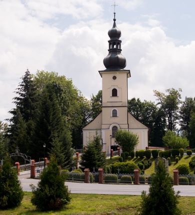 Zdjęcie nr 10: Kościół, zwrócony w kierunku południowo-wschodnim, jest budowlą murowaną z cegły i kamienia, na rzucie podłużnym. Długość bryły wynosi 35, a szerokość 16 metrów. Korpus nawowy jest trójnawowy i czteroprzęsłowy. Stosunkowo szerokie, jednoprzęsłowe, zamknięte półkoliście prezbiterium flankują dwie przyległości, z których jedna jest zakrystią. Na osi kościoła, w przedłużeniu północnego przęsła jest trójkondygnacyjna wieża, ujęta po bokach piętrowymi lokalnościami, lekko wysunięta przed lico fasady. Od zachodu do korpusu przylega kaplica. Nawy przekryte są sklepieniami kolebkowymi wspartymi na filarach o prostokątnym przekroju oraz pilastrach. Przestrzeń prezbiterium od korpusu nawowego oddziela drewniana balustrada. Artykulację ścian na zewnątrz kościoła stanowią osadzone na niewysokim cokole pojedyncze, a w narożach pary lizen wspierających obiegający bryłę fryz, przerwany w partii wieży oraz gzyms. Do wnętrza prowadzą trzy otwory drzwiowe – jeden w elewacji północnej oraz dwa w północnej ścianie kaplicy bocznej. Dwuskrzydłowe drzwi w fasadzie ujęte są profilowanym portalem zamkniętym łukiem odcinkowym zwieńczonym krzyżem. Po bokach wejścia znajduje się para kropielnic. Powyżej otworu drzwiowego usytuowane jest duże okno w kształcie stojącego prostokąta zamkniętego półkoliście, górą ujęte gzymsem. Na wysokości drugiej kondygnacji znajduje się mniejsze okno, zamknięte półkoliście i ujęte prostym obramieniem z kluczem pośrodku oraz parą guttusów pod parapetem. Podobną formę mają cztery okna w ścianach trzeciej kondygnacji wieży, dodatkowo wpisane są one w prostokątne płyciny zamknięte rodzajem łuku kotarowego. W górnej partii wieży znajdują się cztery, okrągłe otwory okienne ujęte prostymi obramieniami zwieńczonymi kluczem. Wieżę wieńczy hełm z osadzoną na cebulastej kopule latarnią zwieńczoną drugą, mniejszą zakończoną krzyżem. Ściany korpusu nawowego i prezbiterium przeprute są prostokątnymi oknami zamkniętymi półkoliście, ujętymi prostym obramieniem z kluczem pośrodku. Okno na zakończeniu prezbiterium jest zamurowane. Okna wypełnione są witrażami, które w prezbiterium zawierają wizerunki Najświętszego Serca Matki Boskiej po prawej i Najświętszego Serca Jezusa po lewej stronie.  Cykl witraży znajduje się także w oknach korpusu, po prawej stronie są to kolejno przedstawienia Matki Boskiej Bolesnej, Boga Ojca, św. Karola Borromeusza i św. Wojciecha, natomiast po lewej stronie – Męża Boleści, Ducha Świętego, św. Jana z Dukli oraz św. Stanisława. Kształt okien korpusu i prezbiterium został powtórzony w kaplicy bocznej, natomiast otwory okienne usytuowane w południowych ścianach przyległości flankujących prezbiterium są w kształcie stojących prostokątów. Korpus nawowy przekryty jest dachem dwuspadowym, wykonanym z blachy. Na wysokości pierwszego przęsła umieszczona jest wieżyczka na sygnaturkę, także obita blachą. Kolorystyka tynków kościoła utrzymana jest w odcieniu jasnożółtym, cokół jest szary natomiast detal architektoniczny jest biały. Plac przykościelny, z wybrukowanymi alejkami i trawnikiem, otoczony jest ogrodzeniem z murowanych słupków i metalowych segmentów.                    