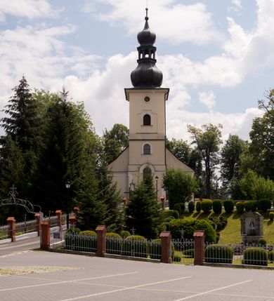 Zdjęcie nr 9: Kościół, zwrócony w kierunku południowo-wschodnim, jest budowlą murowaną z cegły i kamienia, na rzucie podłużnym. Długość bryły wynosi 35, a szerokość 16 metrów. Korpus nawowy jest trójnawowy i czteroprzęsłowy. Stosunkowo szerokie, jednoprzęsłowe, zamknięte półkoliście prezbiterium flankują dwie przyległości, z których jedna jest zakrystią. Na osi kościoła, w przedłużeniu północnego przęsła jest trójkondygnacyjna wieża, ujęta po bokach piętrowymi lokalnościami, lekko wysunięta przed lico fasady. Od zachodu do korpusu przylega kaplica. Nawy przekryte są sklepieniami kolebkowymi wspartymi na filarach o prostokątnym przekroju oraz pilastrach. Przestrzeń prezbiterium od korpusu nawowego oddziela drewniana balustrada. Artykulację ścian na zewnątrz kościoła stanowią osadzone na niewysokim cokole pojedyncze, a w narożach pary lizen wspierających obiegający bryłę fryz, przerwany w partii wieży oraz gzyms. Do wnętrza prowadzą trzy otwory drzwiowe – jeden w elewacji północnej oraz dwa w północnej ścianie kaplicy bocznej. Dwuskrzydłowe drzwi w fasadzie ujęte są profilowanym portalem zamkniętym łukiem odcinkowym zwieńczonym krzyżem. Po bokach wejścia znajduje się para kropielnic. Powyżej otworu drzwiowego usytuowane jest duże okno w kształcie stojącego prostokąta zamkniętego półkoliście, górą ujęte gzymsem. Na wysokości drugiej kondygnacji znajduje się mniejsze okno, zamknięte półkoliście i ujęte prostym obramieniem z kluczem pośrodku oraz parą guttusów pod parapetem. Podobną formę mają cztery okna w ścianach trzeciej kondygnacji wieży, dodatkowo wpisane są one w prostokątne płyciny zamknięte rodzajem łuku kotarowego. W górnej partii wieży znajdują się cztery, okrągłe otwory okienne ujęte prostymi obramieniami zwieńczonymi kluczem. Wieżę wieńczy hełm z osadzoną na cebulastej kopule latarnią zwieńczoną drugą, mniejszą zakończoną krzyżem. Ściany korpusu nawowego i prezbiterium przeprute są prostokątnymi oknami zamkniętymi półkoliście, ujętymi prostym obramieniem z kluczem pośrodku. Okno na zakończeniu prezbiterium jest zamurowane. Okna wypełnione są witrażami, które w prezbiterium zawierają wizerunki Najświętszego Serca Matki Boskiej po prawej i Najświętszego Serca Jezusa po lewej stronie.  Cykl witraży znajduje się także w oknach korpusu, po prawej stronie są to kolejno przedstawienia Matki Boskiej Bolesnej, Boga Ojca, św. Karola Borromeusza i św. Wojciecha, natomiast po lewej stronie – Męża Boleści, Ducha Świętego, św. Jana z Dukli oraz św. Stanisława. Kształt okien korpusu i prezbiterium został powtórzony w kaplicy bocznej, natomiast otwory okienne usytuowane w południowych ścianach przyległości flankujących prezbiterium są w kształcie stojących prostokątów. Korpus nawowy przekryty jest dachem dwuspadowym, wykonanym z blachy. Na wysokości pierwszego przęsła umieszczona jest wieżyczka na sygnaturkę, także obita blachą. Kolorystyka tynków kościoła utrzymana jest w odcieniu jasnożółtym, cokół jest szary natomiast detal architektoniczny jest biały. Plac przykościelny, z wybrukowanymi alejkami i trawnikiem, otoczony jest ogrodzeniem z murowanych słupków i metalowych segmentów.                    