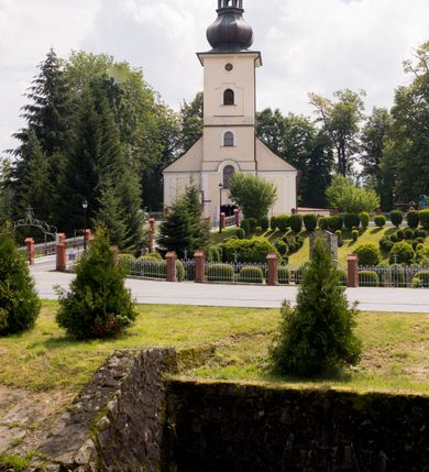 Zdjęcie nr 8: Kościół, zwrócony w kierunku południowo-wschodnim, jest budowlą murowaną z cegły i kamienia, na rzucie podłużnym. Długość bryły wynosi 35, a szerokość 16 metrów. Korpus nawowy jest trójnawowy i czteroprzęsłowy. Stosunkowo szerokie, jednoprzęsłowe, zamknięte półkoliście prezbiterium flankują dwie przyległości, z których jedna jest zakrystią. Na osi kościoła, w przedłużeniu północnego przęsła jest trójkondygnacyjna wieża, ujęta po bokach piętrowymi lokalnościami, lekko wysunięta przed lico fasady. Od zachodu do korpusu przylega kaplica. Nawy przekryte są sklepieniami kolebkowymi wspartymi na filarach o prostokątnym przekroju oraz pilastrach. Przestrzeń prezbiterium od korpusu nawowego oddziela drewniana balustrada. Artykulację ścian na zewnątrz kościoła stanowią osadzone na niewysokim cokole pojedyncze, a w narożach pary lizen wspierających obiegający bryłę fryz, przerwany w partii wieży oraz gzyms. Do wnętrza prowadzą trzy otwory drzwiowe – jeden w elewacji północnej oraz dwa w północnej ścianie kaplicy bocznej. Dwuskrzydłowe drzwi w fasadzie ujęte są profilowanym portalem zamkniętym łukiem odcinkowym zwieńczonym krzyżem. Po bokach wejścia znajduje się para kropielnic. Powyżej otworu drzwiowego usytuowane jest duże okno w kształcie stojącego prostokąta zamkniętego półkoliście, górą ujęte gzymsem. Na wysokości drugiej kondygnacji znajduje się mniejsze okno, zamknięte półkoliście i ujęte prostym obramieniem z kluczem pośrodku oraz parą guttusów pod parapetem. Podobną formę mają cztery okna w ścianach trzeciej kondygnacji wieży, dodatkowo wpisane są one w prostokątne płyciny zamknięte rodzajem łuku kotarowego. W górnej partii wieży znajdują się cztery, okrągłe otwory okienne ujęte prostymi obramieniami zwieńczonymi kluczem. Wieżę wieńczy hełm z osadzoną na cebulastej kopule latarnią zwieńczoną drugą, mniejszą zakończoną krzyżem. Ściany korpusu nawowego i prezbiterium przeprute są prostokątnymi oknami zamkniętymi półkoliście, ujętymi prostym obramieniem z kluczem pośrodku. Okno na zakończeniu prezbiterium jest zamurowane. Okna wypełnione są witrażami, które w prezbiterium zawierają wizerunki Najświętszego Serca Matki Boskiej po prawej i Najświętszego Serca Jezusa po lewej stronie.  Cykl witraży znajduje się także w oknach korpusu, po prawej stronie są to kolejno przedstawienia Matki Boskiej Bolesnej, Boga Ojca, św. Karola Borromeusza i św. Wojciecha, natomiast po lewej stronie – Męża Boleści, Ducha Świętego, św. Jana z Dukli oraz św. Stanisława. Kształt okien korpusu i prezbiterium został powtórzony w kaplicy bocznej, natomiast otwory okienne usytuowane w południowych ścianach przyległości flankujących prezbiterium są w kształcie stojących prostokątów. Korpus nawowy przekryty jest dachem dwuspadowym, wykonanym z blachy. Na wysokości pierwszego przęsła umieszczona jest wieżyczka na sygnaturkę, także obita blachą. Kolorystyka tynków kościoła utrzymana jest w odcieniu jasnożółtym, cokół jest szary natomiast detal architektoniczny jest biały. Plac przykościelny, z wybrukowanymi alejkami i trawnikiem, otoczony jest ogrodzeniem z murowanych słupków i metalowych segmentów.                    