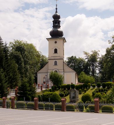 Zdjęcie nr 6: Kościół, zwrócony w kierunku południowo-wschodnim, jest budowlą murowaną z cegły i kamienia, na rzucie podłużnym. Długość bryły wynosi 35, a szerokość 16 metrów. Korpus nawowy jest trójnawowy i czteroprzęsłowy. Stosunkowo szerokie, jednoprzęsłowe, zamknięte półkoliście prezbiterium flankują dwie przyległości, z których jedna jest zakrystią. Na osi kościoła, w przedłużeniu północnego przęsła jest trójkondygnacyjna wieża, ujęta po bokach piętrowymi lokalnościami, lekko wysunięta przed lico fasady. Od zachodu do korpusu przylega kaplica. Nawy przekryte są sklepieniami kolebkowymi wspartymi na filarach o prostokątnym przekroju oraz pilastrach. Przestrzeń prezbiterium od korpusu nawowego oddziela drewniana balustrada. Artykulację ścian na zewnątrz kościoła stanowią osadzone na niewysokim cokole pojedyncze, a w narożach pary lizen wspierających obiegający bryłę fryz, przerwany w partii wieży oraz gzyms. Do wnętrza prowadzą trzy otwory drzwiowe – jeden w elewacji północnej oraz dwa w północnej ścianie kaplicy bocznej. Dwuskrzydłowe drzwi w fasadzie ujęte są profilowanym portalem zamkniętym łukiem odcinkowym zwieńczonym krzyżem. Po bokach wejścia znajduje się para kropielnic. Powyżej otworu drzwiowego usytuowane jest duże okno w kształcie stojącego prostokąta zamkniętego półkoliście, górą ujęte gzymsem. Na wysokości drugiej kondygnacji znajduje się mniejsze okno, zamknięte półkoliście i ujęte prostym obramieniem z kluczem pośrodku oraz parą guttusów pod parapetem. Podobną formę mają cztery okna w ścianach trzeciej kondygnacji wieży, dodatkowo wpisane są one w prostokątne płyciny zamknięte rodzajem łuku kotarowego. W górnej partii wieży znajdują się cztery, okrągłe otwory okienne ujęte prostymi obramieniami zwieńczonymi kluczem. Wieżę wieńczy hełm z osadzoną na cebulastej kopule latarnią zwieńczoną drugą, mniejszą zakończoną krzyżem. Ściany korpusu nawowego i prezbiterium przeprute są prostokątnymi oknami zamkniętymi półkoliście, ujętymi prostym obramieniem z kluczem pośrodku. Okno na zakończeniu prezbiterium jest zamurowane. Okna wypełnione są witrażami, które w prezbiterium zawierają wizerunki Najświętszego Serca Matki Boskiej po prawej i Najświętszego Serca Jezusa po lewej stronie.  Cykl witraży znajduje się także w oknach korpusu, po prawej stronie są to kolejno przedstawienia Matki Boskiej Bolesnej, Boga Ojca, św. Karola Borromeusza i św. Wojciecha, natomiast po lewej stronie – Męża Boleści, Ducha Świętego, św. Jana z Dukli oraz św. Stanisława. Kształt okien korpusu i prezbiterium został powtórzony w kaplicy bocznej, natomiast otwory okienne usytuowane w południowych ścianach przyległości flankujących prezbiterium są w kształcie stojących prostokątów. Korpus nawowy przekryty jest dachem dwuspadowym, wykonanym z blachy. Na wysokości pierwszego przęsła umieszczona jest wieżyczka na sygnaturkę, także obita blachą. Kolorystyka tynków kościoła utrzymana jest w odcieniu jasnożółtym, cokół jest szary natomiast detal architektoniczny jest biały. Plac przykościelny, z wybrukowanymi alejkami i trawnikiem, otoczony jest ogrodzeniem z murowanych słupków i metalowych segmentów.                    