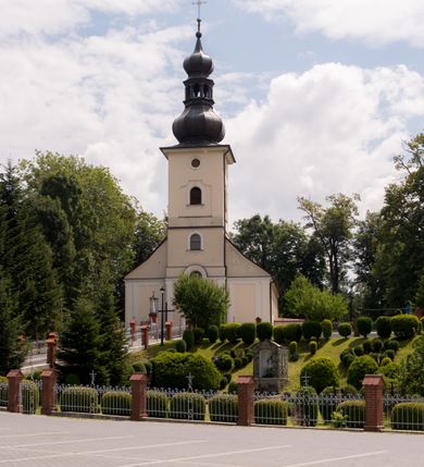 Zdjęcie nr 7: Kościół, zwrócony w kierunku południowo-wschodnim, jest budowlą murowaną z cegły i kamienia, na rzucie podłużnym. Długość bryły wynosi 35, a szerokość 16 metrów. Korpus nawowy jest trójnawowy i czteroprzęsłowy. Stosunkowo szerokie, jednoprzęsłowe, zamknięte półkoliście prezbiterium flankują dwie przyległości, z których jedna jest zakrystią. Na osi kościoła, w przedłużeniu północnego przęsła jest trójkondygnacyjna wieża, ujęta po bokach piętrowymi lokalnościami, lekko wysunięta przed lico fasady. Od zachodu do korpusu przylega kaplica. Nawy przekryte są sklepieniami kolebkowymi wspartymi na filarach o prostokątnym przekroju oraz pilastrach. Przestrzeń prezbiterium od korpusu nawowego oddziela drewniana balustrada. Artykulację ścian na zewnątrz kościoła stanowią osadzone na niewysokim cokole pojedyncze, a w narożach pary lizen wspierających obiegający bryłę fryz, przerwany w partii wieży oraz gzyms. Do wnętrza prowadzą trzy otwory drzwiowe – jeden w elewacji północnej oraz dwa w północnej ścianie kaplicy bocznej. Dwuskrzydłowe drzwi w fasadzie ujęte są profilowanym portalem zamkniętym łukiem odcinkowym zwieńczonym krzyżem. Po bokach wejścia znajduje się para kropielnic. Powyżej otworu drzwiowego usytuowane jest duże okno w kształcie stojącego prostokąta zamkniętego półkoliście, górą ujęte gzymsem. Na wysokości drugiej kondygnacji znajduje się mniejsze okno, zamknięte półkoliście i ujęte prostym obramieniem z kluczem pośrodku oraz parą guttusów pod parapetem. Podobną formę mają cztery okna w ścianach trzeciej kondygnacji wieży, dodatkowo wpisane są one w prostokątne płyciny zamknięte rodzajem łuku kotarowego. W górnej partii wieży znajdują się cztery, okrągłe otwory okienne ujęte prostymi obramieniami zwieńczonymi kluczem. Wieżę wieńczy hełm z osadzoną na cebulastej kopule latarnią zwieńczoną drugą, mniejszą zakończoną krzyżem. Ściany korpusu nawowego i prezbiterium przeprute są prostokątnymi oknami zamkniętymi półkoliście, ujętymi prostym obramieniem z kluczem pośrodku. Okno na zakończeniu prezbiterium jest zamurowane. Okna wypełnione są witrażami, które w prezbiterium zawierają wizerunki Najświętszego Serca Matki Boskiej po prawej i Najświętszego Serca Jezusa po lewej stronie.  Cykl witraży znajduje się także w oknach korpusu, po prawej stronie są to kolejno przedstawienia Matki Boskiej Bolesnej, Boga Ojca, św. Karola Borromeusza i św. Wojciecha, natomiast po lewej stronie – Męża Boleści, Ducha Świętego, św. Jana z Dukli oraz św. Stanisława. Kształt okien korpusu i prezbiterium został powtórzony w kaplicy bocznej, natomiast otwory okienne usytuowane w południowych ścianach przyległości flankujących prezbiterium są w kształcie stojących prostokątów. Korpus nawowy przekryty jest dachem dwuspadowym, wykonanym z blachy. Na wysokości pierwszego przęsła umieszczona jest wieżyczka na sygnaturkę, także obita blachą. Kolorystyka tynków kościoła utrzymana jest w odcieniu jasnożółtym, cokół jest szary natomiast detal architektoniczny jest biały. Plac przykościelny, z wybrukowanymi alejkami i trawnikiem, otoczony jest ogrodzeniem z murowanych słupków i metalowych segmentów.                    