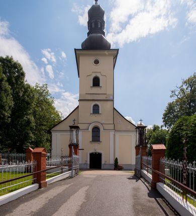 Zdjęcie nr 4: Kościół, zwrócony w kierunku południowo-wschodnim, jest budowlą murowaną z cegły i kamienia, na rzucie podłużnym. Długość bryły wynosi 35, a szerokość 16 metrów. Korpus nawowy jest trójnawowy i czteroprzęsłowy. Stosunkowo szerokie, jednoprzęsłowe, zamknięte półkoliście prezbiterium flankują dwie przyległości, z których jedna jest zakrystią. Na osi kościoła, w przedłużeniu północnego przęsła jest trójkondygnacyjna wieża, ujęta po bokach piętrowymi lokalnościami, lekko wysunięta przed lico fasady. Od zachodu do korpusu przylega kaplica. Nawy przekryte są sklepieniami kolebkowymi wspartymi na filarach o prostokątnym przekroju oraz pilastrach. Przestrzeń prezbiterium od korpusu nawowego oddziela drewniana balustrada. Artykulację ścian na zewnątrz kościoła stanowią osadzone na niewysokim cokole pojedyncze, a w narożach pary lizen wspierających obiegający bryłę fryz, przerwany w partii wieży oraz gzyms. Do wnętrza prowadzą trzy otwory drzwiowe – jeden w elewacji północnej oraz dwa w północnej ścianie kaplicy bocznej. Dwuskrzydłowe drzwi w fasadzie ujęte są profilowanym portalem zamkniętym łukiem odcinkowym zwieńczonym krzyżem. Po bokach wejścia znajduje się para kropielnic. Powyżej otworu drzwiowego usytuowane jest duże okno w kształcie stojącego prostokąta zamkniętego półkoliście, górą ujęte gzymsem. Na wysokości drugiej kondygnacji znajduje się mniejsze okno, zamknięte półkoliście i ujęte prostym obramieniem z kluczem pośrodku oraz parą guttusów pod parapetem. Podobną formę mają cztery okna w ścianach trzeciej kondygnacji wieży, dodatkowo wpisane są one w prostokątne płyciny zamknięte rodzajem łuku kotarowego. W górnej partii wieży znajdują się cztery, okrągłe otwory okienne ujęte prostymi obramieniami zwieńczonymi kluczem. Wieżę wieńczy hełm z osadzoną na cebulastej kopule latarnią zwieńczoną drugą, mniejszą zakończoną krzyżem. Ściany korpusu nawowego i prezbiterium przeprute są prostokątnymi oknami zamkniętymi półkoliście, ujętymi prostym obramieniem z kluczem pośrodku. Okno na zakończeniu prezbiterium jest zamurowane. Okna wypełnione są witrażami, które w prezbiterium zawierają wizerunki Najświętszego Serca Matki Boskiej po prawej i Najświętszego Serca Jezusa po lewej stronie.  Cykl witraży znajduje się także w oknach korpusu, po prawej stronie są to kolejno przedstawienia Matki Boskiej Bolesnej, Boga Ojca, św. Karola Borromeusza i św. Wojciecha, natomiast po lewej stronie – Męża Boleści, Ducha Świętego, św. Jana z Dukli oraz św. Stanisława. Kształt okien korpusu i prezbiterium został powtórzony w kaplicy bocznej, natomiast otwory okienne usytuowane w południowych ścianach przyległości flankujących prezbiterium są w kształcie stojących prostokątów. Korpus nawowy przekryty jest dachem dwuspadowym, wykonanym z blachy. Na wysokości pierwszego przęsła umieszczona jest wieżyczka na sygnaturkę, także obita blachą. Kolorystyka tynków kościoła utrzymana jest w odcieniu jasnożółtym, cokół jest szary natomiast detal architektoniczny jest biały. Plac przykościelny, z wybrukowanymi alejkami i trawnikiem, otoczony jest ogrodzeniem z murowanych słupków i metalowych segmentów.                    