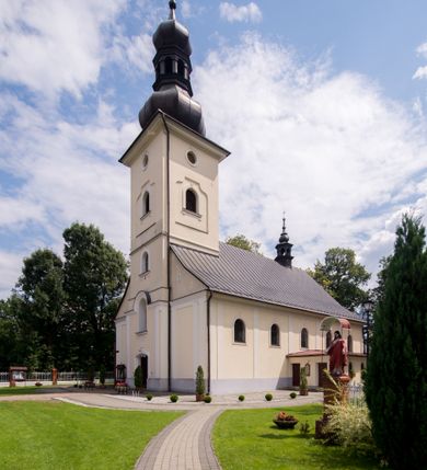 Zdjęcie nr 2: Kościół, zwrócony w kierunku południowo-wschodnim, jest budowlą murowaną z cegły i kamienia, na rzucie podłużnym. Długość bryły wynosi 35, a szerokość 16 metrów. Korpus nawowy jest trójnawowy i czteroprzęsłowy. Stosunkowo szerokie, jednoprzęsłowe, zamknięte półkoliście prezbiterium flankują dwie przyległości, z których jedna jest zakrystią. Na osi kościoła, w przedłużeniu północnego przęsła jest trójkondygnacyjna wieża, ujęta po bokach piętrowymi lokalnościami, lekko wysunięta przed lico fasady. Od zachodu do korpusu przylega kaplica. Nawy przekryte są sklepieniami kolebkowymi wspartymi na filarach o prostokątnym przekroju oraz pilastrach. Przestrzeń prezbiterium od korpusu nawowego oddziela drewniana balustrada. Artykulację ścian na zewnątrz kościoła stanowią osadzone na niewysokim cokole pojedyncze, a w narożach pary lizen wspierających obiegający bryłę fryz, przerwany w partii wieży oraz gzyms. Do wnętrza prowadzą trzy otwory drzwiowe – jeden w elewacji północnej oraz dwa w północnej ścianie kaplicy bocznej. Dwuskrzydłowe drzwi w fasadzie ujęte są profilowanym portalem zamkniętym łukiem odcinkowym zwieńczonym krzyżem. Po bokach wejścia znajduje się para kropielnic. Powyżej otworu drzwiowego usytuowane jest duże okno w kształcie stojącego prostokąta zamkniętego półkoliście, górą ujęte gzymsem. Na wysokości drugiej kondygnacji znajduje się mniejsze okno, zamknięte półkoliście i ujęte prostym obramieniem z kluczem pośrodku oraz parą guttusów pod parapetem. Podobną formę mają cztery okna w ścianach trzeciej kondygnacji wieży, dodatkowo wpisane są one w prostokątne płyciny zamknięte rodzajem łuku kotarowego. W górnej partii wieży znajdują się cztery, okrągłe otwory okienne ujęte prostymi obramieniami zwieńczonymi kluczem. Wieżę wieńczy hełm z osadzoną na cebulastej kopule latarnią zwieńczoną drugą, mniejszą zakończoną krzyżem. Ściany korpusu nawowego i prezbiterium przeprute są prostokątnymi oknami zamkniętymi półkoliście, ujętymi prostym obramieniem z kluczem pośrodku. Okno na zakończeniu prezbiterium jest zamurowane. Okna wypełnione są witrażami, które w prezbiterium zawierają wizerunki Najświętszego Serca Matki Boskiej po prawej i Najświętszego Serca Jezusa po lewej stronie.  Cykl witraży znajduje się także w oknach korpusu, po prawej stronie są to kolejno przedstawienia Matki Boskiej Bolesnej, Boga Ojca, św. Karola Borromeusza i św. Wojciecha, natomiast po lewej stronie – Męża Boleści, Ducha Świętego, św. Jana z Dukli oraz św. Stanisława. Kształt okien korpusu i prezbiterium został powtórzony w kaplicy bocznej, natomiast otwory okienne usytuowane w południowych ścianach przyległości flankujących prezbiterium są w kształcie stojących prostokątów. Korpus nawowy przekryty jest dachem dwuspadowym, wykonanym z blachy. Na wysokości pierwszego przęsła umieszczona jest wieżyczka na sygnaturkę, także obita blachą. Kolorystyka tynków kościoła utrzymana jest w odcieniu jasnożółtym, cokół jest szary natomiast detal architektoniczny jest biały. Plac przykościelny, z wybrukowanymi alejkami i trawnikiem, otoczony jest ogrodzeniem z murowanych słupków i metalowych segmentów.                    