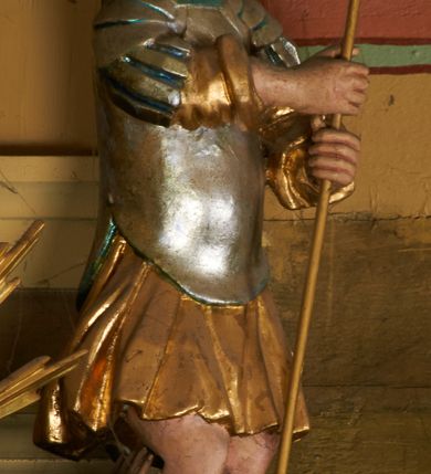 Zdjęcie nr 1: Postać ustawiona jest frontalnie, z głową skierowaną delikatnie w lewo, stoi na lekko ugiętych nogach na leżącej, brązowej postaci diabła. Święty obiema dłońmi trzyma włócznię skierowaną ostrzem w dół. Jego twarz jest pełna, okolona krótkimi włosami. Ubrany jest w strój żołnierza, a więc pozłacaną tunikę sięgającą kolan oraz posrebrzoną zbroję torsową z laserowanymi detalami, na głowie ma srebrzony hełm z pozłoconym pióropuszem, a na nogach wysokie, wiązane i pozłocone sandały. Polichromia naturalistyczna w odsłoniętych partiach ciała.