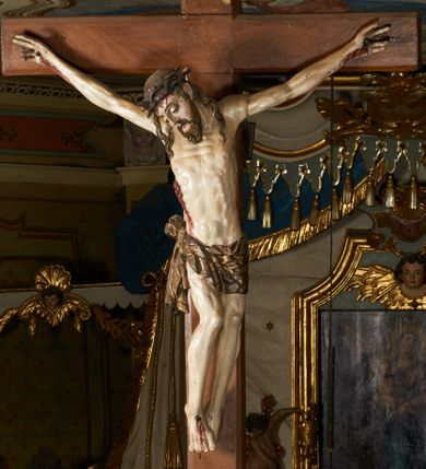 Zdjęcie nr 1: Rzeźba przybita jest trzema gwoździami do prostego krzyża umieszczonego na belce tęczowej. Chrystus ukazany został w typie Cristo morte (umarłego), w lekkim zwisie ciała, o szeroko rozłożonych ramionach przyjmujących kształt delikatnego łuku. Jego głowa opada bezwładnie na prawy bark, nogi ma ugięte w kolanach, skierowane lekko w lewo, w efekcie całe ciało wygina się w pionie w niezbyt wydatny łuk. Stopy skrzyżowane w układzie prawa na lewą. Twarz ma podłużną, o prostym, wąskim nosie, dużych, zamkniętych oczach i niewielkich ustach, okoloną brodą skręconą w dwa pukle oraz pasmami kręconych włosów opadających na ramiona. Na głowie ma koronę cierniową. Jego ciało jest szczupłe, smukłe, o miękko rzeźbionej muskulaturze, silnie podkreślonej w partiach klatki piersiowej, nóg i ramion, z zaznaczonymi śladami męki w postaci broczących rzeźbionymi kroplami krwi ran. Perizonium ma krótkie, ściśle przylegające do ciała, obficie fałdowane zawiązane na prawym biodrze. Polichromia naturalistyczna, perizonium pozłacane. Na pionowej belce krzyża tabliczka z napisem „I.N. / R.I.”. 