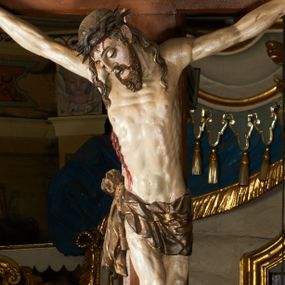 Zdjęcie nr 1: Rzeźba przybita jest trzema gwoździami do prostego krzyża umieszczonego na belce tęczowej. Chrystus ukazany został w typie Cristo morte (umarłego), w lekkim zwisie ciała, o szeroko rozłożonych ramionach przyjmujących kształt delikatnego łuku. Jego głowa opada bezwładnie na prawy bark, nogi ma ugięte w kolanach, skierowane lekko w lewo, w efekcie całe ciało wygina się w pionie w niezbyt wydatny łuk. Stopy skrzyżowane w układzie prawa na lewą. Twarz ma podłużną, o prostym, wąskim nosie, dużych, zamkniętych oczach i niewielkich ustach, okoloną brodą skręconą w dwa pukle oraz pasmami kręconych włosów opadających na ramiona. Na głowie ma koronę cierniową. Jego ciało jest szczupłe, smukłe, o miękko rzeźbionej muskulaturze, silnie podkreślonej w partiach klatki piersiowej, nóg i ramion, z zaznaczonymi śladami męki w postaci broczących rzeźbionymi kroplami krwi ran. Perizonium ma krótkie, ściśle przylegające do ciała, obficie fałdowane zawiązane na prawym biodrze. Polichromia naturalistyczna, perizonium pozłacane. Na pionowej belce krzyża tabliczka z napisem „I.N. / R.I.”. 