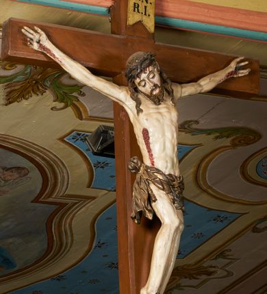Zdjęcie nr 11: Rzeźba przybita jest trzema gwoździami do prostego krzyża umieszczonego na belce tęczowej. Chrystus ukazany został w typie Cristo morte (umarłego), w lekkim zwisie ciała, o szeroko rozłożonych ramionach przyjmujących kształt delikatnego łuku. Jego głowa opada bezwładnie na prawy bark, nogi ma ugięte w kolanach, skierowane lekko w lewo, w efekcie całe ciało wygina się w pionie w niezbyt wydatny łuk. Stopy skrzyżowane w układzie prawa na lewą. Twarz ma podłużną, o prostym, wąskim nosie, dużych, zamkniętych oczach i niewielkich ustach, okoloną brodą skręconą w dwa pukle oraz pasmami kręconych włosów opadających na ramiona. Na głowie ma koronę cierniową. Jego ciało jest szczupłe, smukłe, o miękko rzeźbionej muskulaturze, silnie podkreślonej w partiach klatki piersiowej, nóg i ramion, z zaznaczonymi śladami męki w postaci broczących rzeźbionymi kroplami krwi ran. Perizonium ma krótkie, ściśle przylegające do ciała, obficie fałdowane zawiązane na prawym biodrze. Polichromia naturalistyczna, perizonium pozłacane. Na pionowej belce krzyża tabliczka z napisem „I.N. / R.I.”. 