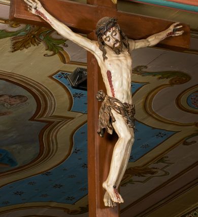 Zdjęcie nr 9: Rzeźba przybita jest trzema gwoździami do prostego krzyża umieszczonego na belce tęczowej. Chrystus ukazany został w typie Cristo morte (umarłego), w lekkim zwisie ciała, o szeroko rozłożonych ramionach przyjmujących kształt delikatnego łuku. Jego głowa opada bezwładnie na prawy bark, nogi ma ugięte w kolanach, skierowane lekko w lewo, w efekcie całe ciało wygina się w pionie w niezbyt wydatny łuk. Stopy skrzyżowane w układzie prawa na lewą. Twarz ma podłużną, o prostym, wąskim nosie, dużych, zamkniętych oczach i niewielkich ustach, okoloną brodą skręconą w dwa pukle oraz pasmami kręconych włosów opadających na ramiona. Na głowie ma koronę cierniową. Jego ciało jest szczupłe, smukłe, o miękko rzeźbionej muskulaturze, silnie podkreślonej w partiach klatki piersiowej, nóg i ramion, z zaznaczonymi śladami męki w postaci broczących rzeźbionymi kroplami krwi ran. Perizonium ma krótkie, ściśle przylegające do ciała, obficie fałdowane zawiązane na prawym biodrze. Polichromia naturalistyczna, perizonium pozłacane. Na pionowej belce krzyża tabliczka z napisem „I.N. / R.I.”. 