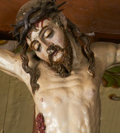 Zdjęcie nr 13: Rzeźba przybita jest trzema gwoździami do prostego krzyża umieszczonego na belce tęczowej. Chrystus ukazany został w typie Cristo morte (umarłego), w lekkim zwisie ciała, o szeroko rozłożonych ramionach przyjmujących kształt delikatnego łuku. Jego głowa opada bezwładnie na prawy bark, nogi ma ugięte w kolanach, skierowane lekko w lewo, w efekcie całe ciało wygina się w pionie w niezbyt wydatny łuk. Stopy skrzyżowane w układzie prawa na lewą. Twarz ma podłużną, o prostym, wąskim nosie, dużych, zamkniętych oczach i niewielkich ustach, okoloną brodą skręconą w dwa pukle oraz pasmami kręconych włosów opadających na ramiona. Na głowie ma koronę cierniową. Jego ciało jest szczupłe, smukłe, o miękko rzeźbionej muskulaturze, silnie podkreślonej w partiach klatki piersiowej, nóg i ramion, z zaznaczonymi śladami męki w postaci broczących rzeźbionymi kroplami krwi ran. Perizonium ma krótkie, ściśle przylegające do ciała, obficie fałdowane zawiązane na prawym biodrze. Polichromia naturalistyczna, perizonium pozłacane. Na pionowej belce krzyża tabliczka z napisem „I.N. / R.I.”. 