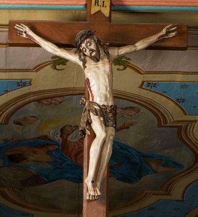 Zdjęcie nr 8: Rzeźba przybita jest trzema gwoździami do prostego krzyża umieszczonego na belce tęczowej. Chrystus ukazany został w typie Cristo morte (umarłego), w lekkim zwisie ciała, o szeroko rozłożonych ramionach przyjmujących kształt delikatnego łuku. Jego głowa opada bezwładnie na prawy bark, nogi ma ugięte w kolanach, skierowane lekko w lewo, w efekcie całe ciało wygina się w pionie w niezbyt wydatny łuk. Stopy skrzyżowane w układzie prawa na lewą. Twarz ma podłużną, o prostym, wąskim nosie, dużych, zamkniętych oczach i niewielkich ustach, okoloną brodą skręconą w dwa pukle oraz pasmami kręconych włosów opadających na ramiona. Na głowie ma koronę cierniową. Jego ciało jest szczupłe, smukłe, o miękko rzeźbionej muskulaturze, silnie podkreślonej w partiach klatki piersiowej, nóg i ramion, z zaznaczonymi śladami męki w postaci broczących rzeźbionymi kroplami krwi ran. Perizonium ma krótkie, ściśle przylegające do ciała, obficie fałdowane zawiązane na prawym biodrze. Polichromia naturalistyczna, perizonium pozłacane. Na pionowej belce krzyża tabliczka z napisem „I.N. / R.I.”. 