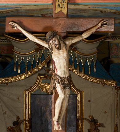 Zdjęcie nr 2: Rzeźba przybita jest trzema gwoździami do prostego krzyża umieszczonego na belce tęczowej. Chrystus ukazany został w typie Cristo morte (umarłego), w lekkim zwisie ciała, o szeroko rozłożonych ramionach przyjmujących kształt delikatnego łuku. Jego głowa opada bezwładnie na prawy bark, nogi ma ugięte w kolanach, skierowane lekko w lewo, w efekcie całe ciało wygina się w pionie w niezbyt wydatny łuk. Stopy skrzyżowane w układzie prawa na lewą. Twarz ma podłużną, o prostym, wąskim nosie, dużych, zamkniętych oczach i niewielkich ustach, okoloną brodą skręconą w dwa pukle oraz pasmami kręconych włosów opadających na ramiona. Na głowie ma koronę cierniową. Jego ciało jest szczupłe, smukłe, o miękko rzeźbionej muskulaturze, silnie podkreślonej w partiach klatki piersiowej, nóg i ramion, z zaznaczonymi śladami męki w postaci broczących rzeźbionymi kroplami krwi ran. Perizonium ma krótkie, ściśle przylegające do ciała, obficie fałdowane zawiązane na prawym biodrze. Polichromia naturalistyczna, perizonium pozłacane. Na pionowej belce krzyża tabliczka z napisem „I.N. / R.I.”. 