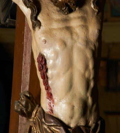 Zdjęcie nr 15: Rzeźba przybita jest trzema gwoździami do prostego krzyża umieszczonego na belce tęczowej. Chrystus ukazany został w typie Cristo morte (umarłego), w lekkim zwisie ciała, o szeroko rozłożonych ramionach przyjmujących kształt delikatnego łuku. Jego głowa opada bezwładnie na prawy bark, nogi ma ugięte w kolanach, skierowane lekko w lewo, w efekcie całe ciało wygina się w pionie w niezbyt wydatny łuk. Stopy skrzyżowane w układzie prawa na lewą. Twarz ma podłużną, o prostym, wąskim nosie, dużych, zamkniętych oczach i niewielkich ustach, okoloną brodą skręconą w dwa pukle oraz pasmami kręconych włosów opadających na ramiona. Na głowie ma koronę cierniową. Jego ciało jest szczupłe, smukłe, o miękko rzeźbionej muskulaturze, silnie podkreślonej w partiach klatki piersiowej, nóg i ramion, z zaznaczonymi śladami męki w postaci broczących rzeźbionymi kroplami krwi ran. Perizonium ma krótkie, ściśle przylegające do ciała, obficie fałdowane zawiązane na prawym biodrze. Polichromia naturalistyczna, perizonium pozłacane. Na pionowej belce krzyża tabliczka z napisem „I.N. / R.I.”. 