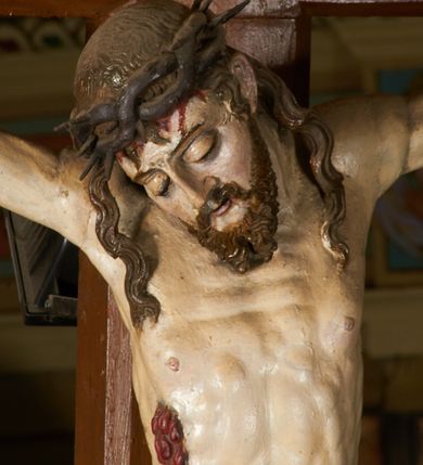 Zdjęcie nr 14: Rzeźba przybita jest trzema gwoździami do prostego krzyża umieszczonego na belce tęczowej. Chrystus ukazany został w typie Cristo morte (umarłego), w lekkim zwisie ciała, o szeroko rozłożonych ramionach przyjmujących kształt delikatnego łuku. Jego głowa opada bezwładnie na prawy bark, nogi ma ugięte w kolanach, skierowane lekko w lewo, w efekcie całe ciało wygina się w pionie w niezbyt wydatny łuk. Stopy skrzyżowane w układzie prawa na lewą. Twarz ma podłużną, o prostym, wąskim nosie, dużych, zamkniętych oczach i niewielkich ustach, okoloną brodą skręconą w dwa pukle oraz pasmami kręconych włosów opadających na ramiona. Na głowie ma koronę cierniową. Jego ciało jest szczupłe, smukłe, o miękko rzeźbionej muskulaturze, silnie podkreślonej w partiach klatki piersiowej, nóg i ramion, z zaznaczonymi śladami męki w postaci broczących rzeźbionymi kroplami krwi ran. Perizonium ma krótkie, ściśle przylegające do ciała, obficie fałdowane zawiązane na prawym biodrze. Polichromia naturalistyczna, perizonium pozłacane. Na pionowej belce krzyża tabliczka z napisem „I.N. / R.I.”. 