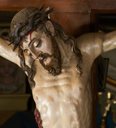 Zdjęcie nr 12: Rzeźba przybita jest trzema gwoździami do prostego krzyża umieszczonego na belce tęczowej. Chrystus ukazany został w typie Cristo morte (umarłego), w lekkim zwisie ciała, o szeroko rozłożonych ramionach przyjmujących kształt delikatnego łuku. Jego głowa opada bezwładnie na prawy bark, nogi ma ugięte w kolanach, skierowane lekko w lewo, w efekcie całe ciało wygina się w pionie w niezbyt wydatny łuk. Stopy skrzyżowane w układzie prawa na lewą. Twarz ma podłużną, o prostym, wąskim nosie, dużych, zamkniętych oczach i niewielkich ustach, okoloną brodą skręconą w dwa pukle oraz pasmami kręconych włosów opadających na ramiona. Na głowie ma koronę cierniową. Jego ciało jest szczupłe, smukłe, o miękko rzeźbionej muskulaturze, silnie podkreślonej w partiach klatki piersiowej, nóg i ramion, z zaznaczonymi śladami męki w postaci broczących rzeźbionymi kroplami krwi ran. Perizonium ma krótkie, ściśle przylegające do ciała, obficie fałdowane zawiązane na prawym biodrze. Polichromia naturalistyczna, perizonium pozłacane. Na pionowej belce krzyża tabliczka z napisem „I.N. / R.I.”. 