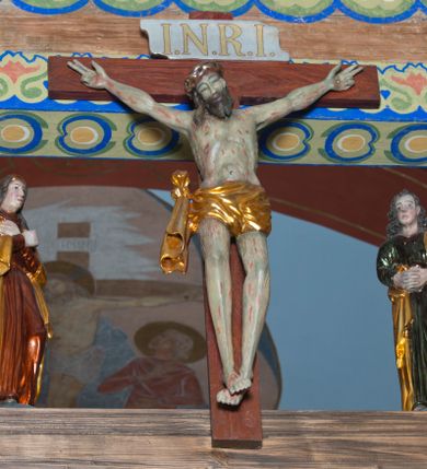 Zdjęcie nr 1: Grupa na tęczy składa się z na wpół ludowo rzeźbionego centralnie krucyfiksu z martwym Chrystusem, po bokach którego stoi pełnorzeźbiona Matka Boska Bolesna i św. Jan Ewangelista. Zbawiciel do krzyża został przymocowany trzema gwoździami. Jezus został ukazany jako martwy z zamkniętymi oczami i głową opadającą na piersi. Jego postać jest wysmukła z nienaturalnie potraktowaną muskulaturą korpusu. Ciało jest wychudzone o zapadłej klatce piersiowej z podkreśloną linią żeber i mostkiem. Z prawej strony widoczny jest przebity bok. Nieznacznie rozchylone nogi są sztywne w kolanach. Prawa stopa założona jest na lewą. Pociągła twarz Chrystusa została miękko opracowana bez szczególnej dbałości o szczegóły anatomiczne. Chrystus ma zamknięte oczy, prosty nos oraz lekko otwarte usta otoczone ciemnobrązowym zarostem, przechodzącym w brodę nieznacznie rozdwojoną na końcu. Jego ciemnobrązowe włosy z lekkim puklowaniem opadają pasmem z prawej strony na ramię, a z lewej na kark, odsłaniając wydatną małżowinę lewego ucha. Chrystus ma na głowie srebrzoną laserunkowo, cierniową koronę, spod której na czoło spływają krople krwi. Krew widoczna jest również na jego rękach, torsie i nogach. Charakterystycznie rzeźbione są nieproporcjonalnie duże dłonie o dwóch zgiętych palcach. Biodra Zbawiciela przepasane są złoconym, krótkim perizonium w formie chusty zawiązanej na duży węzeł z festonem opadającym wzdłuż prawego boku. Ponad głową Chrystusa zawieszony jest titulus w formie tabliczki o faliście wycinanych brzegach ze złotym napisem: „INRI”. 
Stojąca na lekkim wzgórku Matka Boska Bolesna ukazana została w ujęciu trzy czwarte w lewo w nieznacznym kontrapoście. Zgięte w łokciach ręce o nieproporcjonalnie dużych dłoniach ma skrzyżowane na piersi. Maria ma podłużną twarz pozbawioną cech szczególnych. Jej głowę osłania złocona chusta opadająca na ramiona, spod której widoczne są pasma brązowych włosów. Ubrana jest w długą, ciemnoochrową suknię, malowaną laserunkiem, przepasaną w tali oraz płaszcz narzucony na ramiona. Spod sukni widać czubki stóp. 
Święty Jan Ewangelista został zwrócony trzy czwarte w prawo w lekkim kontrapoście. Stojąc na niewielkim wzgórku, trzyma na wysokości brzucha splecione nieproporcjonalnie duże dłonie. Święty ma pociągłą twarz okoloną półdługimi, mocno kręconymi włosami, których pasma z lewej strony odsłaniają wydatną małżowinę uszną. Bardzo mocno zaakcentowano jego krtań. Apostoł ubrany jest w długą tunikę z charakterystycznym kołnierzykiem, malowaną laserunkiem na ciemnooliwkowo oraz złocony płaszcz narzucony na lewe ramię, którego połę zdaje się przyciskać do pasa prawą ręką. Spod tuniki widoczne są czubki stóp. 
Na zachowanym fragmencie oryginalnej belki tęczowej widnieje czerwona inskrypcja: „[...]IT ET IN HONOREN SANCTE MARIE / MAGDALE”. 

