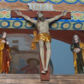 Zdjęcie nr 1: Grupa na tęczy składa się z na wpół ludowo rzeźbionego centralnie krucyfiksu z martwym Chrystusem, po bokach którego stoi pełnorzeźbiona Matka Boska Bolesna i św. Jan Ewangelista. Zbawiciel do krzyża został przymocowany trzema gwoździami. Jezus został ukazany jako martwy z zamkniętymi oczami i głową opadającą na piersi. Jego postać jest wysmukła z nienaturalnie potraktowaną muskulaturą korpusu. Ciało jest wychudzone o zapadłej klatce piersiowej z podkreśloną linią żeber i mostkiem. Z prawej strony widoczny jest przebity bok. Nieznacznie rozchylone nogi są sztywne w kolanach. Prawa stopa założona jest na lewą. Pociągła twarz Chrystusa została miękko opracowana bez szczególnej dbałości o szczegóły anatomiczne. Chrystus ma zamknięte oczy, prosty nos oraz lekko otwarte usta otoczone ciemnobrązowym zarostem, przechodzącym w brodę nieznacznie rozdwojoną na końcu. Jego ciemnobrązowe włosy z lekkim puklowaniem opadają pasmem z prawej strony na ramię, a z lewej na kark, odsłaniając wydatną małżowinę lewego ucha. Chrystus ma na głowie srebrzoną laserunkowo, cierniową koronę, spod której na czoło spływają krople krwi. Krew widoczna jest również na jego rękach, torsie i nogach. Charakterystycznie rzeźbione są nieproporcjonalnie duże dłonie o dwóch zgiętych palcach. Biodra Zbawiciela przepasane są złoconym, krótkim perizonium w formie chusty zawiązanej na duży węzeł z festonem opadającym wzdłuż prawego boku. Ponad głową Chrystusa zawieszony jest titulus w formie tabliczki o faliście wycinanych brzegach ze złotym napisem: „INRI”. 
Stojąca na lekkim wzgórku Matka Boska Bolesna ukazana została w ujęciu trzy czwarte w lewo w nieznacznym kontrapoście. Zgięte w łokciach ręce o nieproporcjonalnie dużych dłoniach ma skrzyżowane na piersi. Maria ma podłużną twarz pozbawioną cech szczególnych. Jej głowę osłania złocona chusta opadająca na ramiona, spod której widoczne są pasma brązowych włosów. Ubrana jest w długą, ciemnoochrową suknię, malowaną laserunkiem, przepasaną w tali oraz płaszcz narzucony na ramiona. Spod sukni widać czubki stóp. 
Święty Jan Ewangelista został zwrócony trzy czwarte w prawo w lekkim kontrapoście. Stojąc na niewielkim wzgórku, trzyma na wysokości brzucha splecione nieproporcjonalnie duże dłonie. Święty ma pociągłą twarz okoloną półdługimi, mocno kręconymi włosami, których pasma z lewej strony odsłaniają wydatną małżowinę uszną. Bardzo mocno zaakcentowano jego krtań. Apostoł ubrany jest w długą tunikę z charakterystycznym kołnierzykiem, malowaną laserunkiem na ciemnooliwkowo oraz złocony płaszcz narzucony na lewe ramię, którego połę zdaje się przyciskać do pasa prawą ręką. Spod tuniki widoczne są czubki stóp. 
Na zachowanym fragmencie oryginalnej belki tęczowej widnieje czerwona inskrypcja: „[...]IT ET IN HONOREN SANCTE MARIE / MAGDALE”. 

