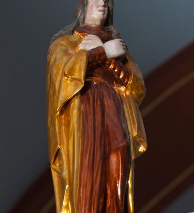 Zdjęcie nr 8: Grupa na tęczy składa się z na wpół ludowo rzeźbionego centralnie krucyfiksu z martwym Chrystusem, po bokach którego stoi pełnorzeźbiona Matka Boska Bolesna i św. Jan Ewangelista. Zbawiciel do krzyża został przymocowany trzema gwoździami. Jezus został ukazany jako martwy z zamkniętymi oczami i głową opadającą na piersi. Jego postać jest wysmukła z nienaturalnie potraktowaną muskulaturą korpusu. Ciało jest wychudzone o zapadłej klatce piersiowej z podkreśloną linią żeber i mostkiem. Z prawej strony widoczny jest przebity bok. Nieznacznie rozchylone nogi są sztywne w kolanach. Prawa stopa założona jest na lewą. Pociągła twarz Chrystusa została miękko opracowana bez szczególnej dbałości o szczegóły anatomiczne. Chrystus ma zamknięte oczy, prosty nos oraz lekko otwarte usta otoczone ciemnobrązowym zarostem, przechodzącym w brodę nieznacznie rozdwojoną na końcu. Jego ciemnobrązowe włosy z lekkim puklowaniem opadają pasmem z prawej strony na ramię, a z lewej na kark, odsłaniając wydatną małżowinę lewego ucha. Chrystus ma na głowie srebrzoną laserunkowo, cierniową koronę, spod której na czoło spływają krople krwi. Krew widoczna jest również na jego rękach, torsie i nogach. Charakterystycznie rzeźbione są nieproporcjonalnie duże dłonie o dwóch zgiętych palcach. Biodra Zbawiciela przepasane są złoconym, krótkim perizonium w formie chusty zawiązanej na duży węzeł z festonem opadającym wzdłuż prawego boku. Ponad głową Chrystusa zawieszony jest titulus w formie tabliczki o faliście wycinanych brzegach ze złotym napisem: „INRI”. 
Stojąca na lekkim wzgórku Matka Boska Bolesna ukazana została w ujęciu trzy czwarte w lewo w nieznacznym kontrapoście. Zgięte w łokciach ręce o nieproporcjonalnie dużych dłoniach ma skrzyżowane na piersi. Maria ma podłużną twarz pozbawioną cech szczególnych. Jej głowę osłania złocona chusta opadająca na ramiona, spod której widoczne są pasma brązowych włosów. Ubrana jest w długą, ciemnoochrową suknię, malowaną laserunkiem, przepasaną w tali oraz płaszcz narzucony na ramiona. Spod sukni widać czubki stóp. 
Święty Jan Ewangelista został zwrócony trzy czwarte w prawo w lekkim kontrapoście. Stojąc na niewielkim wzgórku, trzyma na wysokości brzucha splecione nieproporcjonalnie duże dłonie. Święty ma pociągłą twarz okoloną półdługimi, mocno kręconymi włosami, których pasma z lewej strony odsłaniają wydatną małżowinę uszną. Bardzo mocno zaakcentowano jego krtań. Apostoł ubrany jest w długą tunikę z charakterystycznym kołnierzykiem, malowaną laserunkiem na ciemnooliwkowo oraz złocony płaszcz narzucony na lewe ramię, którego połę zdaje się przyciskać do pasa prawą ręką. Spod tuniki widoczne są czubki stóp. 
Na zachowanym fragmencie oryginalnej belki tęczowej widnieje czerwona inskrypcja: „[...]IT ET IN HONOREN SANCTE MARIE / MAGDALE”. 
