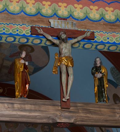 Zdjęcie nr 2: Grupa na tęczy składa się z na wpół ludowo rzeźbionego centralnie krucyfiksu z martwym Chrystusem, po bokach którego stoi pełnorzeźbiona Matka Boska Bolesna i św. Jan Ewangelista. Zbawiciel do krzyża został przymocowany trzema gwoździami. Jezus został ukazany jako martwy z zamkniętymi oczami i głową opadającą na piersi. Jego postać jest wysmukła z nienaturalnie potraktowaną muskulaturą korpusu. Ciało jest wychudzone o zapadłej klatce piersiowej z podkreśloną linią żeber i mostkiem. Z prawej strony widoczny jest przebity bok. Nieznacznie rozchylone nogi są sztywne w kolanach. Prawa stopa założona jest na lewą. Pociągła twarz Chrystusa została miękko opracowana bez szczególnej dbałości o szczegóły anatomiczne. Chrystus ma zamknięte oczy, prosty nos oraz lekko otwarte usta otoczone ciemnobrązowym zarostem, przechodzącym w brodę nieznacznie rozdwojoną na końcu. Jego ciemnobrązowe włosy z lekkim puklowaniem opadają pasmem z prawej strony na ramię, a z lewej na kark, odsłaniając wydatną małżowinę lewego ucha. Chrystus ma na głowie srebrzoną laserunkowo, cierniową koronę, spod której na czoło spływają krople krwi. Krew widoczna jest również na jego rękach, torsie i nogach. Charakterystycznie rzeźbione są nieproporcjonalnie duże dłonie o dwóch zgiętych palcach. Biodra Zbawiciela przepasane są złoconym, krótkim perizonium w formie chusty zawiązanej na duży węzeł z festonem opadającym wzdłuż prawego boku. Ponad głową Chrystusa zawieszony jest titulus w formie tabliczki o faliście wycinanych brzegach ze złotym napisem: „INRI”. 
Stojąca na lekkim wzgórku Matka Boska Bolesna ukazana została w ujęciu trzy czwarte w lewo w nieznacznym kontrapoście. Zgięte w łokciach ręce o nieproporcjonalnie dużych dłoniach ma skrzyżowane na piersi. Maria ma podłużną twarz pozbawioną cech szczególnych. Jej głowę osłania złocona chusta opadająca na ramiona, spod której widoczne są pasma brązowych włosów. Ubrana jest w długą, ciemnoochrową suknię, malowaną laserunkiem, przepasaną w tali oraz płaszcz narzucony na ramiona. Spod sukni widać czubki stóp. 
Święty Jan Ewangelista został zwrócony trzy czwarte w prawo w lekkim kontrapoście. Stojąc na niewielkim wzgórku, trzyma na wysokości brzucha splecione nieproporcjonalnie duże dłonie. Święty ma pociągłą twarz okoloną półdługimi, mocno kręconymi włosami, których pasma z lewej strony odsłaniają wydatną małżowinę uszną. Bardzo mocno zaakcentowano jego krtań. Apostoł ubrany jest w długą tunikę z charakterystycznym kołnierzykiem, malowaną laserunkiem na ciemnooliwkowo oraz złocony płaszcz narzucony na lewe ramię, którego połę zdaje się przyciskać do pasa prawą ręką. Spod tuniki widoczne są czubki stóp. 
Na zachowanym fragmencie oryginalnej belki tęczowej widnieje czerwona inskrypcja: „[...]IT ET IN HONOREN SANCTE MARIE / MAGDALE”. 
