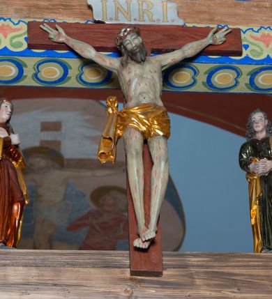 Zdjęcie nr 4: Grupa na tęczy składa się z na wpół ludowo rzeźbionego centralnie krucyfiksu z martwym Chrystusem, po bokach którego stoi pełnorzeźbiona Matka Boska Bolesna i św. Jan Ewangelista. Zbawiciel do krzyża został przymocowany trzema gwoździami. Jezus został ukazany jako martwy z zamkniętymi oczami i głową opadającą na piersi. Jego postać jest wysmukła z nienaturalnie potraktowaną muskulaturą korpusu. Ciało jest wychudzone o zapadłej klatce piersiowej z podkreśloną linią żeber i mostkiem. Z prawej strony widoczny jest przebity bok. Nieznacznie rozchylone nogi są sztywne w kolanach. Prawa stopa założona jest na lewą. Pociągła twarz Chrystusa została miękko opracowana bez szczególnej dbałości o szczegóły anatomiczne. Chrystus ma zamknięte oczy, prosty nos oraz lekko otwarte usta otoczone ciemnobrązowym zarostem, przechodzącym w brodę nieznacznie rozdwojoną na końcu. Jego ciemnobrązowe włosy z lekkim puklowaniem opadają pasmem z prawej strony na ramię, a z lewej na kark, odsłaniając wydatną małżowinę lewego ucha. Chrystus ma na głowie srebrzoną laserunkowo, cierniową koronę, spod której na czoło spływają krople krwi. Krew widoczna jest również na jego rękach, torsie i nogach. Charakterystycznie rzeźbione są nieproporcjonalnie duże dłonie o dwóch zgiętych palcach. Biodra Zbawiciela przepasane są złoconym, krótkim perizonium w formie chusty zawiązanej na duży węzeł z festonem opadającym wzdłuż prawego boku. Ponad głową Chrystusa zawieszony jest titulus w formie tabliczki o faliście wycinanych brzegach ze złotym napisem: „INRI”. 
Stojąca na lekkim wzgórku Matka Boska Bolesna ukazana została w ujęciu trzy czwarte w lewo w nieznacznym kontrapoście. Zgięte w łokciach ręce o nieproporcjonalnie dużych dłoniach ma skrzyżowane na piersi. Maria ma podłużną twarz pozbawioną cech szczególnych. Jej głowę osłania złocona chusta opadająca na ramiona, spod której widoczne są pasma brązowych włosów. Ubrana jest w długą, ciemnoochrową suknię, malowaną laserunkiem, przepasaną w tali oraz płaszcz narzucony na ramiona. Spod sukni widać czubki stóp. 
Święty Jan Ewangelista został zwrócony trzy czwarte w prawo w lekkim kontrapoście. Stojąc na niewielkim wzgórku, trzyma na wysokości brzucha splecione nieproporcjonalnie duże dłonie. Święty ma pociągłą twarz okoloną półdługimi, mocno kręconymi włosami, których pasma z lewej strony odsłaniają wydatną małżowinę uszną. Bardzo mocno zaakcentowano jego krtań. Apostoł ubrany jest w długą tunikę z charakterystycznym kołnierzykiem, malowaną laserunkiem na ciemnooliwkowo oraz złocony płaszcz narzucony na lewe ramię, którego połę zdaje się przyciskać do pasa prawą ręką. Spod tuniki widoczne są czubki stóp. 
Na zachowanym fragmencie oryginalnej belki tęczowej widnieje czerwona inskrypcja: „[...]IT ET IN HONOREN SANCTE MARIE / MAGDALE”. 
