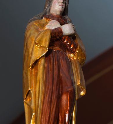 Zdjęcie nr 9: Grupa na tęczy składa się z na wpół ludowo rzeźbionego centralnie krucyfiksu z martwym Chrystusem, po bokach którego stoi pełnorzeźbiona Matka Boska Bolesna i św. Jan Ewangelista. Zbawiciel do krzyża został przymocowany trzema gwoździami. Jezus został ukazany jako martwy z zamkniętymi oczami i głową opadającą na piersi. Jego postać jest wysmukła z nienaturalnie potraktowaną muskulaturą korpusu. Ciało jest wychudzone o zapadłej klatce piersiowej z podkreśloną linią żeber i mostkiem. Z prawej strony widoczny jest przebity bok. Nieznacznie rozchylone nogi są sztywne w kolanach. Prawa stopa założona jest na lewą. Pociągła twarz Chrystusa została miękko opracowana bez szczególnej dbałości o szczegóły anatomiczne. Chrystus ma zamknięte oczy, prosty nos oraz lekko otwarte usta otoczone ciemnobrązowym zarostem, przechodzącym w brodę nieznacznie rozdwojoną na końcu. Jego ciemnobrązowe włosy z lekkim puklowaniem opadają pasmem z prawej strony na ramię, a z lewej na kark, odsłaniając wydatną małżowinę lewego ucha. Chrystus ma na głowie srebrzoną laserunkowo, cierniową koronę, spod której na czoło spływają krople krwi. Krew widoczna jest również na jego rękach, torsie i nogach. Charakterystycznie rzeźbione są nieproporcjonalnie duże dłonie o dwóch zgiętych palcach. Biodra Zbawiciela przepasane są złoconym, krótkim perizonium w formie chusty zawiązanej na duży węzeł z festonem opadającym wzdłuż prawego boku. Ponad głową Chrystusa zawieszony jest titulus w formie tabliczki o faliście wycinanych brzegach ze złotym napisem: „INRI”. 
Stojąca na lekkim wzgórku Matka Boska Bolesna ukazana została w ujęciu trzy czwarte w lewo w nieznacznym kontrapoście. Zgięte w łokciach ręce o nieproporcjonalnie dużych dłoniach ma skrzyżowane na piersi. Maria ma podłużną twarz pozbawioną cech szczególnych. Jej głowę osłania złocona chusta opadająca na ramiona, spod której widoczne są pasma brązowych włosów. Ubrana jest w długą, ciemnoochrową suknię, malowaną laserunkiem, przepasaną w tali oraz płaszcz narzucony na ramiona. Spod sukni widać czubki stóp. 
Święty Jan Ewangelista został zwrócony trzy czwarte w prawo w lekkim kontrapoście. Stojąc na niewielkim wzgórku, trzyma na wysokości brzucha splecione nieproporcjonalnie duże dłonie. Święty ma pociągłą twarz okoloną półdługimi, mocno kręconymi włosami, których pasma z lewej strony odsłaniają wydatną małżowinę uszną. Bardzo mocno zaakcentowano jego krtań. Apostoł ubrany jest w długą tunikę z charakterystycznym kołnierzykiem, malowaną laserunkiem na ciemnooliwkowo oraz złocony płaszcz narzucony na lewe ramię, którego połę zdaje się przyciskać do pasa prawą ręką. Spod tuniki widoczne są czubki stóp. 
Na zachowanym fragmencie oryginalnej belki tęczowej widnieje czerwona inskrypcja: „[...]IT ET IN HONOREN SANCTE MARIE / MAGDALE”. 
