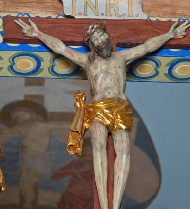 Zdjęcie nr 5: Grupa na tęczy składa się z na wpół ludowo rzeźbionego centralnie krucyfiksu z martwym Chrystusem, po bokach którego stoi pełnorzeźbiona Matka Boska Bolesna i św. Jan Ewangelista. Zbawiciel do krzyża został przymocowany trzema gwoździami. Jezus został ukazany jako martwy z zamkniętymi oczami i głową opadającą na piersi. Jego postać jest wysmukła z nienaturalnie potraktowaną muskulaturą korpusu. Ciało jest wychudzone o zapadłej klatce piersiowej z podkreśloną linią żeber i mostkiem. Z prawej strony widoczny jest przebity bok. Nieznacznie rozchylone nogi są sztywne w kolanach. Prawa stopa założona jest na lewą. Pociągła twarz Chrystusa została miękko opracowana bez szczególnej dbałości o szczegóły anatomiczne. Chrystus ma zamknięte oczy, prosty nos oraz lekko otwarte usta otoczone ciemnobrązowym zarostem, przechodzącym w brodę nieznacznie rozdwojoną na końcu. Jego ciemnobrązowe włosy z lekkim puklowaniem opadają pasmem z prawej strony na ramię, a z lewej na kark, odsłaniając wydatną małżowinę lewego ucha. Chrystus ma na głowie srebrzoną laserunkowo, cierniową koronę, spod której na czoło spływają krople krwi. Krew widoczna jest również na jego rękach, torsie i nogach. Charakterystycznie rzeźbione są nieproporcjonalnie duże dłonie o dwóch zgiętych palcach. Biodra Zbawiciela przepasane są złoconym, krótkim perizonium w formie chusty zawiązanej na duży węzeł z festonem opadającym wzdłuż prawego boku. Ponad głową Chrystusa zawieszony jest titulus w formie tabliczki o faliście wycinanych brzegach ze złotym napisem: „INRI”. 
Stojąca na lekkim wzgórku Matka Boska Bolesna ukazana została w ujęciu trzy czwarte w lewo w nieznacznym kontrapoście. Zgięte w łokciach ręce o nieproporcjonalnie dużych dłoniach ma skrzyżowane na piersi. Maria ma podłużną twarz pozbawioną cech szczególnych. Jej głowę osłania złocona chusta opadająca na ramiona, spod której widoczne są pasma brązowych włosów. Ubrana jest w długą, ciemnoochrową suknię, malowaną laserunkiem, przepasaną w tali oraz płaszcz narzucony na ramiona. Spod sukni widać czubki stóp. 
Święty Jan Ewangelista został zwrócony trzy czwarte w prawo w lekkim kontrapoście. Stojąc na niewielkim wzgórku, trzyma na wysokości brzucha splecione nieproporcjonalnie duże dłonie. Święty ma pociągłą twarz okoloną półdługimi, mocno kręconymi włosami, których pasma z lewej strony odsłaniają wydatną małżowinę uszną. Bardzo mocno zaakcentowano jego krtań. Apostoł ubrany jest w długą tunikę z charakterystycznym kołnierzykiem, malowaną laserunkiem na ciemnooliwkowo oraz złocony płaszcz narzucony na lewe ramię, którego połę zdaje się przyciskać do pasa prawą ręką. Spod tuniki widoczne są czubki stóp. 
Na zachowanym fragmencie oryginalnej belki tęczowej widnieje czerwona inskrypcja: „[...]IT ET IN HONOREN SANCTE MARIE / MAGDALE”. 
