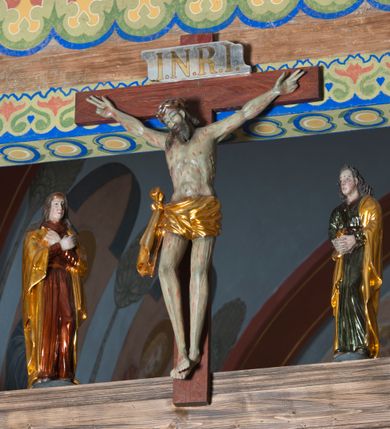 Zdjęcie nr 3: Grupa na tęczy składa się z na wpół ludowo rzeźbionego centralnie krucyfiksu z martwym Chrystusem, po bokach którego stoi pełnorzeźbiona Matka Boska Bolesna i św. Jan Ewangelista. Zbawiciel do krzyża został przymocowany trzema gwoździami. Jezus został ukazany jako martwy z zamkniętymi oczami i głową opadającą na piersi. Jego postać jest wysmukła z nienaturalnie potraktowaną muskulaturą korpusu. Ciało jest wychudzone o zapadłej klatce piersiowej z podkreśloną linią żeber i mostkiem. Z prawej strony widoczny jest przebity bok. Nieznacznie rozchylone nogi są sztywne w kolanach. Prawa stopa założona jest na lewą. Pociągła twarz Chrystusa została miękko opracowana bez szczególnej dbałości o szczegóły anatomiczne. Chrystus ma zamknięte oczy, prosty nos oraz lekko otwarte usta otoczone ciemnobrązowym zarostem, przechodzącym w brodę nieznacznie rozdwojoną na końcu. Jego ciemnobrązowe włosy z lekkim puklowaniem opadają pasmem z prawej strony na ramię, a z lewej na kark, odsłaniając wydatną małżowinę lewego ucha. Chrystus ma na głowie srebrzoną laserunkowo, cierniową koronę, spod której na czoło spływają krople krwi. Krew widoczna jest również na jego rękach, torsie i nogach. Charakterystycznie rzeźbione są nieproporcjonalnie duże dłonie o dwóch zgiętych palcach. Biodra Zbawiciela przepasane są złoconym, krótkim perizonium w formie chusty zawiązanej na duży węzeł z festonem opadającym wzdłuż prawego boku. Ponad głową Chrystusa zawieszony jest titulus w formie tabliczki o faliście wycinanych brzegach ze złotym napisem: „INRI”. 
Stojąca na lekkim wzgórku Matka Boska Bolesna ukazana została w ujęciu trzy czwarte w lewo w nieznacznym kontrapoście. Zgięte w łokciach ręce o nieproporcjonalnie dużych dłoniach ma skrzyżowane na piersi. Maria ma podłużną twarz pozbawioną cech szczególnych. Jej głowę osłania złocona chusta opadająca na ramiona, spod której widoczne są pasma brązowych włosów. Ubrana jest w długą, ciemnoochrową suknię, malowaną laserunkiem, przepasaną w tali oraz płaszcz narzucony na ramiona. Spod sukni widać czubki stóp. 
Święty Jan Ewangelista został zwrócony trzy czwarte w prawo w lekkim kontrapoście. Stojąc na niewielkim wzgórku, trzyma na wysokości brzucha splecione nieproporcjonalnie duże dłonie. Święty ma pociągłą twarz okoloną półdługimi, mocno kręconymi włosami, których pasma z lewej strony odsłaniają wydatną małżowinę uszną. Bardzo mocno zaakcentowano jego krtań. Apostoł ubrany jest w długą tunikę z charakterystycznym kołnierzykiem, malowaną laserunkiem na ciemnooliwkowo oraz złocony płaszcz narzucony na lewe ramię, którego połę zdaje się przyciskać do pasa prawą ręką. Spod tuniki widoczne są czubki stóp. 
Na zachowanym fragmencie oryginalnej belki tęczowej widnieje czerwona inskrypcja: „[...]IT ET IN HONOREN SANCTE MARIE / MAGDALE”. 
