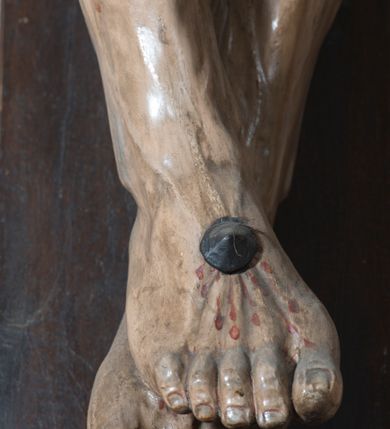 Zdjęcie nr 9: Na dużym krzyżu z ciemnego drewna zawieszono polichromowaną figurę martwego Chrystusa o szeroko rozpostartych ramionach. Ciało Zbawiciela do krzyża jest przymocowane za pomocą trzech gwoździ. On sam został ukazany jako martwy z zamkniętymi oczami i nieznacznie słaniającą się głową. Jego postać jest wysmukła z nienaturalnie potraktowaną muskulaturą korpusu. Wyraźnie zaznaczono naprężone żyły na całym ciele, co ma sprawiać wrażenie jego autentyczności. Mocno również zaznaczono krwawiącą wybroczynę z przebitego prawego boku. Ciało jest wychudzone o zapadłej klatce piersiowej z silnie podkreśloną linią żeber i mostkiem. Nogi są lekko ugięte w kolanach. Prawa stopa założona jest na lewą. Płaskie, niezbyt szczegółowe opracowanie pociągłej twarzy, o charakterystycznym mięsistym dużym nosie, zamkniętych oczach i nieznacznie rozchylonych ustach sprawia, że w figurze trudno dopatrzeć się ekspresji odpowiadającej dramatowi ukrzyżowania. Głowę otaczają brązowe włosy opadające puklami na kark oraz na prawe ramię. Twarz Jezusa okala brązowy zarost z charakterystycznym przedzieleniem na brodzie. Chrystus ma na głowie złoconą, cierniową koronę, spod której na czoło spływają krople krwi. Biodra Zbawiciela przepasane są złoconym, krótkim perizonium w formie chusty zawiązanej na duży węzeł z rozwianym festonem przy prawym boku. Jest ono przewiązane sznurem widocznym fragmentarycznie z prawej strony na tle odsłoniętego prawego biodra. Tkanina perizonium jest mocno drapowana w partii podbrzusza, gdzie widać wywinięty fragment draperii układający się w fałdę misową. Ponad głową Chrystusa na pionowej belce krzyża zawieszono rozwiany titulus z napisem „IN / RI”.
