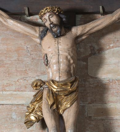Zdjęcie nr 1: Na dużym krzyżu z ciemnego drewna zawieszono polichromowaną figurę martwego Chrystusa o szeroko rozpostartych ramionach. Ciało Zbawiciela do krzyża jest przymocowane za pomocą trzech gwoździ. On sam został ukazany jako martwy z zamkniętymi oczami i nieznacznie słaniającą się głową. Jego postać jest wysmukła z nienaturalnie potraktowaną muskulaturą korpusu. Wyraźnie zaznaczono naprężone żyły na całym ciele, co ma sprawiać wrażenie jego autentyczności. Mocno również zaznaczono krwawiącą wybroczynę z przebitego prawego boku. Ciało jest wychudzone o zapadłej klatce piersiowej z silnie podkreśloną linią żeber i mostkiem. Nogi są lekko ugięte w kolanach. Prawa stopa założona jest na lewą. Płaskie, niezbyt szczegółowe opracowanie pociągłej twarzy, o charakterystycznym mięsistym dużym nosie, zamkniętych oczach i nieznacznie rozchylonych ustach sprawia, że w figurze trudno dopatrzeć się ekspresji odpowiadającej dramatowi ukrzyżowania. Głowę otaczają brązowe włosy opadające puklami na kark oraz na prawe ramię. Twarz Jezusa okala brązowy zarost z charakterystycznym przedzieleniem na brodzie. Chrystus ma na głowie złoconą, cierniową koronę, spod której na czoło spływają krople krwi. Biodra Zbawiciela przepasane są złoconym, krótkim perizonium w formie chusty zawiązanej na duży węzeł z rozwianym festonem przy prawym boku. Jest ono przewiązane sznurem widocznym fragmentarycznie z prawej strony na tle odsłoniętego prawego biodra. Tkanina perizonium jest mocno drapowana w partii podbrzusza, gdzie widać wywinięty fragment draperii układający się w fałdę misową. Ponad głową Chrystusa na pionowej belce krzyża zawieszono rozwiany titulus z napisem „IN / RI”.
