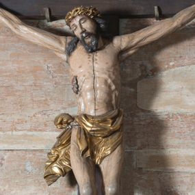 Zdjęcie nr 1: Na dużym krzyżu z ciemnego drewna zawieszono polichromowaną figurę martwego Chrystusa o szeroko rozpostartych ramionach. Ciało Zbawiciela do krzyża jest przymocowane za pomocą trzech gwoździ. On sam został ukazany jako martwy z zamkniętymi oczami i nieznacznie słaniającą się głową. Jego postać jest wysmukła z nienaturalnie potraktowaną muskulaturą korpusu. Wyraźnie zaznaczono naprężone żyły na całym ciele, co ma sprawiać wrażenie jego autentyczności. Mocno również zaznaczono krwawiącą wybroczynę z przebitego prawego boku. Ciało jest wychudzone o zapadłej klatce piersiowej z silnie podkreśloną linią żeber i mostkiem. Nogi są lekko ugięte w kolanach. Prawa stopa założona jest na lewą. Płaskie, niezbyt szczegółowe opracowanie pociągłej twarzy, o charakterystycznym mięsistym dużym nosie, zamkniętych oczach i nieznacznie rozchylonych ustach sprawia, że w figurze trudno dopatrzeć się ekspresji odpowiadającej dramatowi ukrzyżowania. Głowę otaczają brązowe włosy opadające puklami na kark oraz na prawe ramię. Twarz Jezusa okala brązowy zarost z charakterystycznym przedzieleniem na brodzie. Chrystus ma na głowie złoconą, cierniową koronę, spod której na czoło spływają krople krwi. Biodra Zbawiciela przepasane są złoconym, krótkim perizonium w formie chusty zawiązanej na duży węzeł z rozwianym festonem przy prawym boku. Jest ono przewiązane sznurem widocznym fragmentarycznie z prawej strony na tle odsłoniętego prawego biodra. Tkanina perizonium jest mocno drapowana w partii podbrzusza, gdzie widać wywinięty fragment draperii układający się w fałdę misową. Ponad głową Chrystusa na pionowej belce krzyża zawieszono rozwiany titulus z napisem „IN / RI”.
