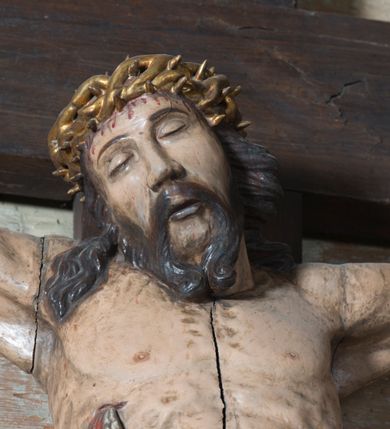 Zdjęcie nr 5: Na dużym krzyżu z ciemnego drewna zawieszono polichromowaną figurę martwego Chrystusa o szeroko rozpostartych ramionach. Ciało Zbawiciela do krzyża jest przymocowane za pomocą trzech gwoździ. On sam został ukazany jako martwy z zamkniętymi oczami i nieznacznie słaniającą się głową. Jego postać jest wysmukła z nienaturalnie potraktowaną muskulaturą korpusu. Wyraźnie zaznaczono naprężone żyły na całym ciele, co ma sprawiać wrażenie jego autentyczności. Mocno również zaznaczono krwawiącą wybroczynę z przebitego prawego boku. Ciało jest wychudzone o zapadłej klatce piersiowej z silnie podkreśloną linią żeber i mostkiem. Nogi są lekko ugięte w kolanach. Prawa stopa założona jest na lewą. Płaskie, niezbyt szczegółowe opracowanie pociągłej twarzy, o charakterystycznym mięsistym dużym nosie, zamkniętych oczach i nieznacznie rozchylonych ustach sprawia, że w figurze trudno dopatrzeć się ekspresji odpowiadającej dramatowi ukrzyżowania. Głowę otaczają brązowe włosy opadające puklami na kark oraz na prawe ramię. Twarz Jezusa okala brązowy zarost z charakterystycznym przedzieleniem na brodzie. Chrystus ma na głowie złoconą, cierniową koronę, spod której na czoło spływają krople krwi. Biodra Zbawiciela przepasane są złoconym, krótkim perizonium w formie chusty zawiązanej na duży węzeł z rozwianym festonem przy prawym boku. Jest ono przewiązane sznurem widocznym fragmentarycznie z prawej strony na tle odsłoniętego prawego biodra. Tkanina perizonium jest mocno drapowana w partii podbrzusza, gdzie widać wywinięty fragment draperii układający się w fałdę misową. Ponad głową Chrystusa na pionowej belce krzyża zawieszono rozwiany titulus z napisem „IN / RI”.
