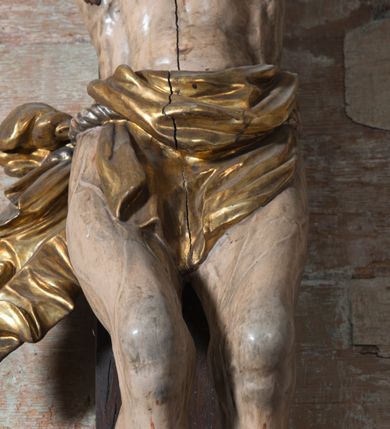 Zdjęcie nr 7: Na dużym krzyżu z ciemnego drewna zawieszono polichromowaną figurę martwego Chrystusa o szeroko rozpostartych ramionach. Ciało Zbawiciela do krzyża jest przymocowane za pomocą trzech gwoździ. On sam został ukazany jako martwy z zamkniętymi oczami i nieznacznie słaniającą się głową. Jego postać jest wysmukła z nienaturalnie potraktowaną muskulaturą korpusu. Wyraźnie zaznaczono naprężone żyły na całym ciele, co ma sprawiać wrażenie jego autentyczności. Mocno również zaznaczono krwawiącą wybroczynę z przebitego prawego boku. Ciało jest wychudzone o zapadłej klatce piersiowej z silnie podkreśloną linią żeber i mostkiem. Nogi są lekko ugięte w kolanach. Prawa stopa założona jest na lewą. Płaskie, niezbyt szczegółowe opracowanie pociągłej twarzy, o charakterystycznym mięsistym dużym nosie, zamkniętych oczach i nieznacznie rozchylonych ustach sprawia, że w figurze trudno dopatrzeć się ekspresji odpowiadającej dramatowi ukrzyżowania. Głowę otaczają brązowe włosy opadające puklami na kark oraz na prawe ramię. Twarz Jezusa okala brązowy zarost z charakterystycznym przedzieleniem na brodzie. Chrystus ma na głowie złoconą, cierniową koronę, spod której na czoło spływają krople krwi. Biodra Zbawiciela przepasane są złoconym, krótkim perizonium w formie chusty zawiązanej na duży węzeł z rozwianym festonem przy prawym boku. Jest ono przewiązane sznurem widocznym fragmentarycznie z prawej strony na tle odsłoniętego prawego biodra. Tkanina perizonium jest mocno drapowana w partii podbrzusza, gdzie widać wywinięty fragment draperii układający się w fałdę misową. Ponad głową Chrystusa na pionowej belce krzyża zawieszono rozwiany titulus z napisem „IN / RI”.
