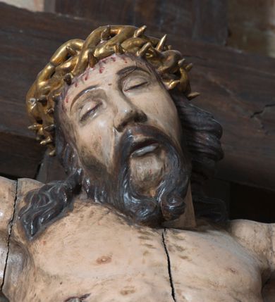 Zdjęcie nr 6: Na dużym krzyżu z ciemnego drewna zawieszono polichromowaną figurę martwego Chrystusa o szeroko rozpostartych ramionach. Ciało Zbawiciela do krzyża jest przymocowane za pomocą trzech gwoździ. On sam został ukazany jako martwy z zamkniętymi oczami i nieznacznie słaniającą się głową. Jego postać jest wysmukła z nienaturalnie potraktowaną muskulaturą korpusu. Wyraźnie zaznaczono naprężone żyły na całym ciele, co ma sprawiać wrażenie jego autentyczności. Mocno również zaznaczono krwawiącą wybroczynę z przebitego prawego boku. Ciało jest wychudzone o zapadłej klatce piersiowej z silnie podkreśloną linią żeber i mostkiem. Nogi są lekko ugięte w kolanach. Prawa stopa założona jest na lewą. Płaskie, niezbyt szczegółowe opracowanie pociągłej twarzy, o charakterystycznym mięsistym dużym nosie, zamkniętych oczach i nieznacznie rozchylonych ustach sprawia, że w figurze trudno dopatrzeć się ekspresji odpowiadającej dramatowi ukrzyżowania. Głowę otaczają brązowe włosy opadające puklami na kark oraz na prawe ramię. Twarz Jezusa okala brązowy zarost z charakterystycznym przedzieleniem na brodzie. Chrystus ma na głowie złoconą, cierniową koronę, spod której na czoło spływają krople krwi. Biodra Zbawiciela przepasane są złoconym, krótkim perizonium w formie chusty zawiązanej na duży węzeł z rozwianym festonem przy prawym boku. Jest ono przewiązane sznurem widocznym fragmentarycznie z prawej strony na tle odsłoniętego prawego biodra. Tkanina perizonium jest mocno drapowana w partii podbrzusza, gdzie widać wywinięty fragment draperii układający się w fałdę misową. Ponad głową Chrystusa na pionowej belce krzyża zawieszono rozwiany titulus z napisem „IN / RI”.
