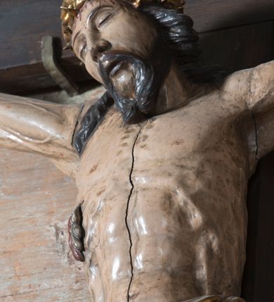Zdjęcie nr 3: Na dużym krzyżu z ciemnego drewna zawieszono polichromowaną figurę martwego Chrystusa o szeroko rozpostartych ramionach. Ciało Zbawiciela do krzyża jest przymocowane za pomocą trzech gwoździ. On sam został ukazany jako martwy z zamkniętymi oczami i nieznacznie słaniającą się głową. Jego postać jest wysmukła z nienaturalnie potraktowaną muskulaturą korpusu. Wyraźnie zaznaczono naprężone żyły na całym ciele, co ma sprawiać wrażenie jego autentyczności. Mocno również zaznaczono krwawiącą wybroczynę z przebitego prawego boku. Ciało jest wychudzone o zapadłej klatce piersiowej z silnie podkreśloną linią żeber i mostkiem. Nogi są lekko ugięte w kolanach. Prawa stopa założona jest na lewą. Płaskie, niezbyt szczegółowe opracowanie pociągłej twarzy, o charakterystycznym mięsistym dużym nosie, zamkniętych oczach i nieznacznie rozchylonych ustach sprawia, że w figurze trudno dopatrzeć się ekspresji odpowiadającej dramatowi ukrzyżowania. Głowę otaczają brązowe włosy opadające puklami na kark oraz na prawe ramię. Twarz Jezusa okala brązowy zarost z charakterystycznym przedzieleniem na brodzie. Chrystus ma na głowie złoconą, cierniową koronę, spod której na czoło spływają krople krwi. Biodra Zbawiciela przepasane są złoconym, krótkim perizonium w formie chusty zawiązanej na duży węzeł z rozwianym festonem przy prawym boku. Jest ono przewiązane sznurem widocznym fragmentarycznie z prawej strony na tle odsłoniętego prawego biodra. Tkanina perizonium jest mocno drapowana w partii podbrzusza, gdzie widać wywinięty fragment draperii układający się w fałdę misową. Ponad głową Chrystusa na pionowej belce krzyża zawieszono rozwiany titulus z napisem „IN / RI”.
