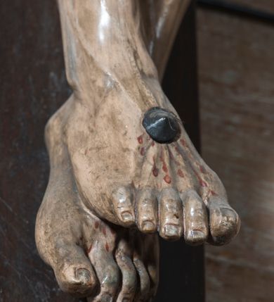 Zdjęcie nr 10: Na dużym krzyżu z ciemnego drewna zawieszono polichromowaną figurę martwego Chrystusa o szeroko rozpostartych ramionach. Ciało Zbawiciela do krzyża jest przymocowane za pomocą trzech gwoździ. On sam został ukazany jako martwy z zamkniętymi oczami i nieznacznie słaniającą się głową. Jego postać jest wysmukła z nienaturalnie potraktowaną muskulaturą korpusu. Wyraźnie zaznaczono naprężone żyły na całym ciele, co ma sprawiać wrażenie jego autentyczności. Mocno również zaznaczono krwawiącą wybroczynę z przebitego prawego boku. Ciało jest wychudzone o zapadłej klatce piersiowej z silnie podkreśloną linią żeber i mostkiem. Nogi są lekko ugięte w kolanach. Prawa stopa założona jest na lewą. Płaskie, niezbyt szczegółowe opracowanie pociągłej twarzy, o charakterystycznym mięsistym dużym nosie, zamkniętych oczach i nieznacznie rozchylonych ustach sprawia, że w figurze trudno dopatrzeć się ekspresji odpowiadającej dramatowi ukrzyżowania. Głowę otaczają brązowe włosy opadające puklami na kark oraz na prawe ramię. Twarz Jezusa okala brązowy zarost z charakterystycznym przedzieleniem na brodzie. Chrystus ma na głowie złoconą, cierniową koronę, spod której na czoło spływają krople krwi. Biodra Zbawiciela przepasane są złoconym, krótkim perizonium w formie chusty zawiązanej na duży węzeł z rozwianym festonem przy prawym boku. Jest ono przewiązane sznurem widocznym fragmentarycznie z prawej strony na tle odsłoniętego prawego biodra. Tkanina perizonium jest mocno drapowana w partii podbrzusza, gdzie widać wywinięty fragment draperii układający się w fałdę misową. Ponad głową Chrystusa na pionowej belce krzyża zawieszono rozwiany titulus z napisem „IN / RI”.
