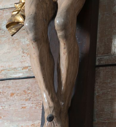 Zdjęcie nr 8: Na dużym krzyżu z ciemnego drewna zawieszono polichromowaną figurę martwego Chrystusa o szeroko rozpostartych ramionach. Ciało Zbawiciela do krzyża jest przymocowane za pomocą trzech gwoździ. On sam został ukazany jako martwy z zamkniętymi oczami i nieznacznie słaniającą się głową. Jego postać jest wysmukła z nienaturalnie potraktowaną muskulaturą korpusu. Wyraźnie zaznaczono naprężone żyły na całym ciele, co ma sprawiać wrażenie jego autentyczności. Mocno również zaznaczono krwawiącą wybroczynę z przebitego prawego boku. Ciało jest wychudzone o zapadłej klatce piersiowej z silnie podkreśloną linią żeber i mostkiem. Nogi są lekko ugięte w kolanach. Prawa stopa założona jest na lewą. Płaskie, niezbyt szczegółowe opracowanie pociągłej twarzy, o charakterystycznym mięsistym dużym nosie, zamkniętych oczach i nieznacznie rozchylonych ustach sprawia, że w figurze trudno dopatrzeć się ekspresji odpowiadającej dramatowi ukrzyżowania. Głowę otaczają brązowe włosy opadające puklami na kark oraz na prawe ramię. Twarz Jezusa okala brązowy zarost z charakterystycznym przedzieleniem na brodzie. Chrystus ma na głowie złoconą, cierniową koronę, spod której na czoło spływają krople krwi. Biodra Zbawiciela przepasane są złoconym, krótkim perizonium w formie chusty zawiązanej na duży węzeł z rozwianym festonem przy prawym boku. Jest ono przewiązane sznurem widocznym fragmentarycznie z prawej strony na tle odsłoniętego prawego biodra. Tkanina perizonium jest mocno drapowana w partii podbrzusza, gdzie widać wywinięty fragment draperii układający się w fałdę misową. Ponad głową Chrystusa na pionowej belce krzyża zawieszono rozwiany titulus z napisem „IN / RI”.
