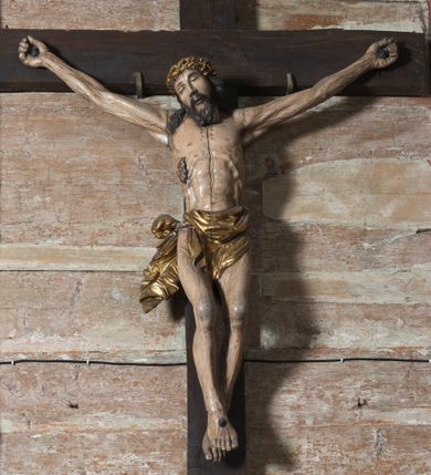 Zdjęcie nr 2: Na dużym krzyżu z ciemnego drewna zawieszono polichromowaną figurę martwego Chrystusa o szeroko rozpostartych ramionach. Ciało Zbawiciela do krzyża jest przymocowane za pomocą trzech gwoździ. On sam został ukazany jako martwy z zamkniętymi oczami i nieznacznie słaniającą się głową. Jego postać jest wysmukła z nienaturalnie potraktowaną muskulaturą korpusu. Wyraźnie zaznaczono naprężone żyły na całym ciele, co ma sprawiać wrażenie jego autentyczności. Mocno również zaznaczono krwawiącą wybroczynę z przebitego prawego boku. Ciało jest wychudzone o zapadłej klatce piersiowej z silnie podkreśloną linią żeber i mostkiem. Nogi są lekko ugięte w kolanach. Prawa stopa założona jest na lewą. Płaskie, niezbyt szczegółowe opracowanie pociągłej twarzy, o charakterystycznym mięsistym dużym nosie, zamkniętych oczach i nieznacznie rozchylonych ustach sprawia, że w figurze trudno dopatrzeć się ekspresji odpowiadającej dramatowi ukrzyżowania. Głowę otaczają brązowe włosy opadające puklami na kark oraz na prawe ramię. Twarz Jezusa okala brązowy zarost z charakterystycznym przedzieleniem na brodzie. Chrystus ma na głowie złoconą, cierniową koronę, spod której na czoło spływają krople krwi. Biodra Zbawiciela przepasane są złoconym, krótkim perizonium w formie chusty zawiązanej na duży węzeł z rozwianym festonem przy prawym boku. Jest ono przewiązane sznurem widocznym fragmentarycznie z prawej strony na tle odsłoniętego prawego biodra. Tkanina perizonium jest mocno drapowana w partii podbrzusza, gdzie widać wywinięty fragment draperii układający się w fałdę misową. Ponad głową Chrystusa na pionowej belce krzyża zawieszono rozwiany titulus z napisem „IN / RI”.
