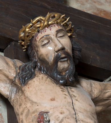 Zdjęcie nr 4: Na dużym krzyżu z ciemnego drewna zawieszono polichromowaną figurę martwego Chrystusa o szeroko rozpostartych ramionach. Ciało Zbawiciela do krzyża jest przymocowane za pomocą trzech gwoździ. On sam został ukazany jako martwy z zamkniętymi oczami i nieznacznie słaniającą się głową. Jego postać jest wysmukła z nienaturalnie potraktowaną muskulaturą korpusu. Wyraźnie zaznaczono naprężone żyły na całym ciele, co ma sprawiać wrażenie jego autentyczności. Mocno również zaznaczono krwawiącą wybroczynę z przebitego prawego boku. Ciało jest wychudzone o zapadłej klatce piersiowej z silnie podkreśloną linią żeber i mostkiem. Nogi są lekko ugięte w kolanach. Prawa stopa założona jest na lewą. Płaskie, niezbyt szczegółowe opracowanie pociągłej twarzy, o charakterystycznym mięsistym dużym nosie, zamkniętych oczach i nieznacznie rozchylonych ustach sprawia, że w figurze trudno dopatrzeć się ekspresji odpowiadającej dramatowi ukrzyżowania. Głowę otaczają brązowe włosy opadające puklami na kark oraz na prawe ramię. Twarz Jezusa okala brązowy zarost z charakterystycznym przedzieleniem na brodzie. Chrystus ma na głowie złoconą, cierniową koronę, spod której na czoło spływają krople krwi. Biodra Zbawiciela przepasane są złoconym, krótkim perizonium w formie chusty zawiązanej na duży węzeł z rozwianym festonem przy prawym boku. Jest ono przewiązane sznurem widocznym fragmentarycznie z prawej strony na tle odsłoniętego prawego biodra. Tkanina perizonium jest mocno drapowana w partii podbrzusza, gdzie widać wywinięty fragment draperii układający się w fałdę misową. Ponad głową Chrystusa na pionowej belce krzyża zawieszono rozwiany titulus z napisem „IN / RI”.
