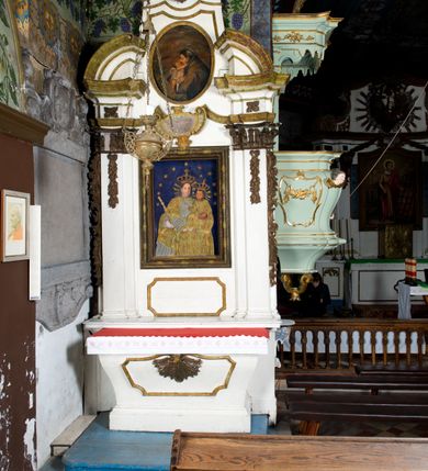 Zdjęcie nr 2: Przyścienne retabulum wznosi się nad sarkofagowym stipesie z drewnianą mensą z kamiennym portatylem, ustawowym na cokole, wspartym na drewnianym podeście. Stipes zwęża się ku dołowi. Od frontu zdobi go fantazyjne obramienie utworzone z profilowanej listwy przechodzącej na środku w płaskorzeźbiony ornament muszlowy. Ponad ołtarzem wznosi się jednoosiowe i jednokondygnacyjne retabulum ze zwieńczeniem ustawione na wysokim cokole. Nastawę artykułuje para zdwojonych pilastrów o korynckich głowicach z trzonami dekorowanymi zawieszonymi na wstęgach festonach owocowo-kwiatowych. Pilastry dźwigają imposty o wyłamanym wklęśle gzymsie, oraz przerwany półkolisty przyczółek. Imposty połączone zostały architrawem nad polem głównym, przyjmującym kształt odwróconego łuku odcinkowego, dekorowanego muszlowym kartuszem z napisem: „ORA PRO NOBIS / SANCTA DEI / GENITRIX”. W polu środkowym umieszczono obraz Matki Boskiej z Dzieciątkiem w formie stojącego prostokąta ujęty profilowaną ramą. Poniżej obrazu znalazła się ramka w formie leżącego prostokąta o wklęsłych bokach. Zwieńczenie uzyskało formę stojącego prostokąta o wklęsłych bokach zamknięty całującymi się wolutami i muszlą. W polu zwieńczenia umieszczono owalny obraz św. Antoniego Padewskiego, ujęty profilowaną ramą. Ponad nim znajdują się dekoracyjne, profilowane listewki układające się w fantazyjne kształty. Po bokach retabulum usytuowano zredukowane uszaki w formie wąskich, ażurowych wici roślinnych. Strukturę nastawy polichromowano na biało, ornamentyka i profile pierwotnie były złocone.
