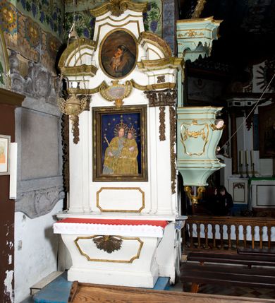 Zdjęcie nr 3: Przyścienne retabulum wznosi się nad sarkofagowym stipesie z drewnianą mensą z kamiennym portatylem, ustawowym na cokole, wspartym na drewnianym podeście. Stipes zwęża się ku dołowi. Od frontu zdobi go fantazyjne obramienie utworzone z profilowanej listwy przechodzącej na środku w płaskorzeźbiony ornament muszlowy. Ponad ołtarzem wznosi się jednoosiowe i jednokondygnacyjne retabulum ze zwieńczeniem ustawione na wysokim cokole. Nastawę artykułuje para zdwojonych pilastrów o korynckich głowicach z trzonami dekorowanymi zawieszonymi na wstęgach festonach owocowo-kwiatowych. Pilastry dźwigają imposty o wyłamanym wklęśle gzymsie, oraz przerwany półkolisty przyczółek. Imposty połączone zostały architrawem nad polem głównym, przyjmującym kształt odwróconego łuku odcinkowego, dekorowanego muszlowym kartuszem z napisem: „ORA PRO NOBIS / SANCTA DEI / GENITRIX”. W polu środkowym umieszczono obraz Matki Boskiej z Dzieciątkiem w formie stojącego prostokąta ujęty profilowaną ramą. Poniżej obrazu znalazła się ramka w formie leżącego prostokąta o wklęsłych bokach. Zwieńczenie uzyskało formę stojącego prostokąta o wklęsłych bokach zamknięty całującymi się wolutami i muszlą. W polu zwieńczenia umieszczono owalny obraz św. Antoniego Padewskiego, ujęty profilowaną ramą. Ponad nim znajdują się dekoracyjne, profilowane listewki układające się w fantazyjne kształty. Po bokach retabulum usytuowano zredukowane uszaki w formie wąskich, ażurowych wici roślinnych. Strukturę nastawy polichromowano na biało, ornamentyka i profile pierwotnie były złocone.