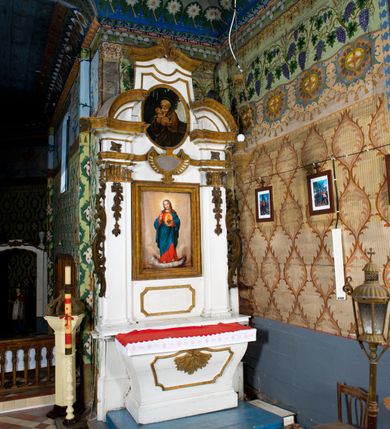Zdjęcie nr 3: Przyścienne retabulum wznosi się nad sarkofagowym stipesie z drewnianą mensą z kamiennym portatylem, ustawowym na cokole, wspartym na drewnianym podeście. Stipes zwęża się ku dołowi. Od frontu zdobi go fantazyjne obramienie utworzone z profilowanej listwy przechodzącej na środku w płaskorzeźbiony ornament muszlowy. Ponad ołtarzem wznosi się jednoosiowe i jednokondygnacyjne retabulum ze zwieńczeniem ustawione na wysokim cokole. Nastawę artykułuje para zdwojonych pilastrów o korynckich głowicach z trzonami dekorowanymi zawieszonymi na wstęgach festonach owocowo-kwiatowych. Pilastry dźwigają imposty o wyłamanym wklęśle gzymsie, oraz przerwany półkolisty przyczółek. Imposty połączone zostały architrawem nad polem głównym, przyjmującym kształt odwróconego łuku odcinkowego, dekorowanego muszlowym kartuszem. W polu środkowym umieszczono obraz Najświętszego Serca Pana Jezusa w formie stojącego prostokąta ujęty profilowaną ramą. Poniżej obrazu znalazła się ramka w formie leżącego prostokąta o wklęsłych bokach. Zwieńczenie uzyskało formę stojącego prostokąta o wklęsłych bokach zamknięty całującymi się wolutami i muszlą. W polu zwieńczenia umieszczono owalny obraz św. Józefa, ujęty profilowaną ramą. Ponad nim znajdują się dekoracyjne, profilowane listewki układające się w fantazyjne kształty. Po bokach retabulum usytuowano zredukowane uszaki w formie wąskich, ażurowych wici roślinnych. Strukturę nastawy polichromowano na biało, ornamentyka i profile pierwotnie były złocone.