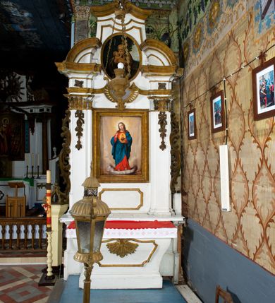 Zdjęcie nr 4: Przyścienne retabulum wznosi się nad sarkofagowym stipesie z drewnianą mensą z kamiennym portatylem, ustawowym na cokole, wspartym na drewnianym podeście. Stipes zwęża się ku dołowi. Od frontu zdobi go fantazyjne obramienie utworzone z profilowanej listwy przechodzącej na środku w płaskorzeźbiony ornament muszlowy. Ponad ołtarzem wznosi się jednoosiowe i jednokondygnacyjne retabulum ze zwieńczeniem ustawione na wysokim cokole. Nastawę artykułuje para zdwojonych pilastrów o korynckich głowicach z trzonami dekorowanymi zawieszonymi na wstęgach festonach owocowo-kwiatowych. Pilastry dźwigają imposty o wyłamanym wklęśle gzymsie, oraz przerwany półkolisty przyczółek. Imposty połączone zostały architrawem nad polem głównym, przyjmującym kształt odwróconego łuku odcinkowego, dekorowanego muszlowym kartuszem. W polu środkowym umieszczono obraz Najświętszego Serca Pana Jezusa w formie stojącego prostokąta ujęty profilowaną ramą. Poniżej obrazu znalazła się ramka w formie leżącego prostokąta o wklęsłych bokach. Zwieńczenie uzyskało formę stojącego prostokąta o wklęsłych bokach zamknięty całującymi się wolutami i muszlą. W polu zwieńczenia umieszczono owalny obraz św. Józefa, ujęty profilowaną ramą. Ponad nim znajdują się dekoracyjne, profilowane listewki układające się w fantazyjne kształty. Po bokach retabulum usytuowano zredukowane uszaki w formie wąskich, ażurowych wici roślinnych. Strukturę nastawy polichromowano na biało, ornamentyka i profile pierwotnie były złocone.