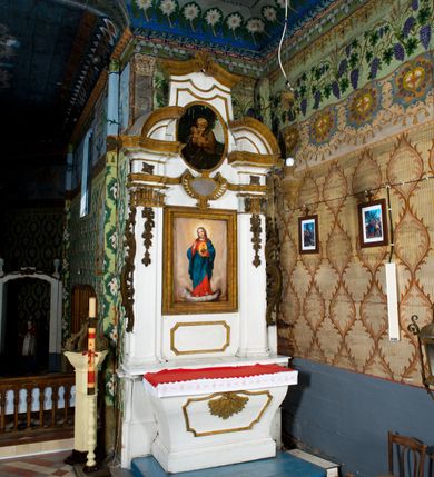 Zdjęcie nr 2: Przyścienne retabulum wznosi się nad sarkofagowym stipesie z drewnianą mensą z kamiennym portatylem, ustawowym na cokole, wspartym na drewnianym podeście. Stipes zwęża się ku dołowi. Od frontu zdobi go fantazyjne obramienie utworzone z profilowanej listwy przechodzącej na środku w płaskorzeźbiony ornament muszlowy. Ponad ołtarzem wznosi się jednoosiowe i jednokondygnacyjne retabulum ze zwieńczeniem ustawione na wysokim cokole. Nastawę artykułuje para zdwojonych pilastrów o korynckich głowicach z trzonami dekorowanymi zawieszonymi na wstęgach festonach owocowo-kwiatowych. Pilastry dźwigają imposty o wyłamanym wklęśle gzymsie, oraz przerwany półkolisty przyczółek. Imposty połączone zostały architrawem nad polem głównym, przyjmującym kształt odwróconego łuku odcinkowego, dekorowanego muszlowym kartuszem. W polu środkowym umieszczono obraz Najświętszego Serca Pana Jezusa w formie stojącego prostokąta ujęty profilowaną ramą. Poniżej obrazu znalazła się ramka w formie leżącego prostokąta o wklęsłych bokach. Zwieńczenie uzyskało formę stojącego prostokąta o wklęsłych bokach zamknięty całującymi się wolutami i muszlą. W polu zwieńczenia umieszczono owalny obraz św. Józefa, ujęty profilowaną ramą. Ponad nim znajdują się dekoracyjne, profilowane listewki układające się w fantazyjne kształty. Po bokach retabulum usytuowano zredukowane uszaki w formie wąskich, ażurowych wici roślinnych. Strukturę nastawy polichromowano na biało, ornamentyka i profile pierwotnie były złocone.