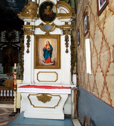 Zdjęcie nr 5: Przyścienne retabulum wznosi się nad sarkofagowym stipesie z drewnianą mensą z kamiennym portatylem, ustawowym na cokole, wspartym na drewnianym podeście. Stipes zwęża się ku dołowi. Od frontu zdobi go fantazyjne obramienie utworzone z profilowanej listwy przechodzącej na środku w płaskorzeźbiony ornament muszlowy. Ponad ołtarzem wznosi się jednoosiowe i jednokondygnacyjne retabulum ze zwieńczeniem ustawione na wysokim cokole. Nastawę artykułuje para zdwojonych pilastrów o korynckich głowicach z trzonami dekorowanymi zawieszonymi na wstęgach festonach owocowo-kwiatowych. Pilastry dźwigają imposty o wyłamanym wklęśle gzymsie, oraz przerwany półkolisty przyczółek. Imposty połączone zostały architrawem nad polem głównym, przyjmującym kształt odwróconego łuku odcinkowego, dekorowanego muszlowym kartuszem. W polu środkowym umieszczono obraz Najświętszego Serca Pana Jezusa w formie stojącego prostokąta ujęty profilowaną ramą. Poniżej obrazu znalazła się ramka w formie leżącego prostokąta o wklęsłych bokach. Zwieńczenie uzyskało formę stojącego prostokąta o wklęsłych bokach zamknięty całującymi się wolutami i muszlą. W polu zwieńczenia umieszczono owalny obraz św. Józefa, ujęty profilowaną ramą. Ponad nim znajdują się dekoracyjne, profilowane listewki układające się w fantazyjne kształty. Po bokach retabulum usytuowano zredukowane uszaki w formie wąskich, ażurowych wici roślinnych. Strukturę nastawy polichromowano na biało, ornamentyka i profile pierwotnie były złocone.