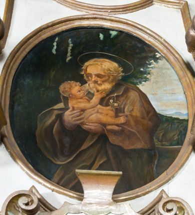Zdjęcie nr 1: Obraz uzyskał kształt stojącego owalu. W centrum kompozycji w dwóch trzecich ukazano stojącego św. Józefa, piastującego Dzieciątko Jezus. Święty został ujęty w trzech czwartych w prawo. W obu rękach uniesionych na wysokości piersi trzyma małego Jezusa. Józefa ukazano jako starego mężczyznę z bujnymi, siwymi, krótkimi włosami i taką samą brodą. Ma wysuszoną twarz o jasnej karnacji, wydatny nos i usta. Jego spojrzenie półprzymknietych oczu skierowane jest na Dzieciątko. Ubrany jest w ciemnozieloną tunikę oraz mocno udrapowany, ciemnobrązowy płaszcz obficie spowijający jego postać. Głowę świętego okala nimb. Nagie Dzieciątko prawą ręką dotyka brody swojego opiekuna. W lewej natomiast ułożonej wzdłuż tułowia trzyma gałązkę z kwiatami. Mały Jezus ma owalną twarz, drobny nos i wydatne usta. W głębi widać sekwencję z drzewami, górzysty pejzaż oraz niebo zakryte białymi obłokami. 