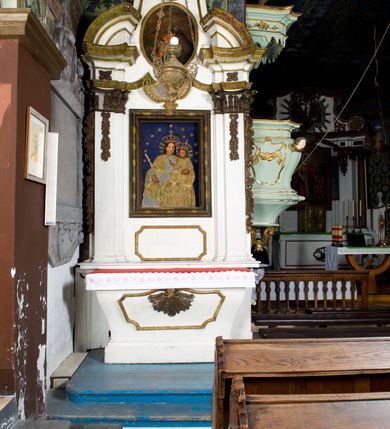 Zdjęcie nr 2: Obraz uzyskał formę stojącego prostokąta. W centrum kompozycji na ciemnoniebieskim tle utworzonym z tkaniny ukazano frontalnie w dwóch trzecich Madonnę z Dzieciątkiem. Obie postacie przykrywa płaskorzeźbiona, drewniana sukienka wotywna z koronami i berłem. Matka Boska na lewym ramieniu trzyma Dzieciątko, a w prawej ręce berło. Głowa Madonny ujęta została w trzech czwartych w lewo. Maria ma pociągłą twarz, wysokie czoło, prosty, długi nos i wydatne usta oraz ciemne oczy o spojrzeniu skierowanym na widza. Jej głowę otaczają ciemne włosy spływające na ramiona. Ubrana jest w luźną, srebrzoną suknię przepasaną w pasie i narzucony na nią złocony płaszcz. Pod szyją ma zawieszony złocony krzyżyk. Dzieciątko Jezus ukazane zostało w trzech czwartych w prawo. Prawą rękę ma uniesioną w geście błogosławieństwa, lewą zaś wspiera na lewej dłoni matki. Jezus ma owalną twarz, wydatny nos i usta oraz szeroko otwarte ciemne oczy o spojrzeniu skierowanym na widza. Na głowach obu postaci otoczonych promieniami widnieją korony zamknięte oraz wieniec z gwiazd.