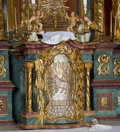 Zdjęcie nr 44: Kamienny ołtarz w jawiszowickim kościele uzyskał kształt prostopadłościanu. Otacza go drewniana obudowa podtrzymująca drewnianą mensę z portatylem. Jego przód zdobi drewniane antepedium ujęte profilowaną ramą z centralnym medalionem z monogramem Marii, otoczonym przez ornament roślinny. Ponad ołtarzem rozpościera się dwukondygnacyjna, architektoniczna nastawa ołtarzowa, flankowana przez dwie bramki. Struktura ołtarza dekorowana jest ciemnozieloną marmoryzacją. Pierwsza kondygnacja retabulum uzyskała trójosiowe założenie wsparte na dwustrefowym cokole, podzielonym płycinami z dekoracją akantową. Artykułują ją dwie pary kolumn o skręconych trzonach i kapitelach kompozytowych. Złocone w całości kolumny podtrzymują belkowanie o wysuniętym gzymsie, wyłamane na ich osi oraz przerwany trójkątny przyczółek. Pole środkowe nastawy wypełnia w całości nisza ujęta profilowaną, złoconą ramą w formie stojącego prostokąta zamkniętego trzema bokami z ośmioboku. W jej wnętrzu znajduje się obraz Matki Boskiej z Dzieciątkiem w typie Matki Boskiej Śnieżnej, zdobiony drewnianą złoconą i srebrzoną sukienką wotywną oraz koronami, a także dwie zasuwy z przedstawieniem św. Marcina, biskupa oraz bł. Salomei. Niszę środkową flankują znajdujące się pomiędzy kolumnami wąskie płyciny zwieńczone konchami. Na ich tle ustawiono figury św. Piotra i św. Pawła. Ponad nimi widoczne są na belkowaniu płaskorzeźbione i złocone girlandy kwiatowe oraz rozety. Na przerwanym przyczółku umieszczono dwie figury siedzących aniołów. Na mensie ołtarza centralnie ustawiono prostopadłościenne tabernakulum w drewnianej obudowie ujęte dwiema kolumienkami kompozytowymi o skręconych trzonach podtrzymujących belkowanie, do którego przymocowano świeczniki na sakramentki. Umieszczony na jego szczycie prosty tron eucharystyczny bez zaplecka flankują dwie figury uskrzydlonych aniołków. Drugą, jednoosiową kondygnację nastawy ujmuje także para złoconych kolumn o skręconych trzonach i kapitelach kompozytowych, ustawionych na cokołach, dźwigających imposty. Pomiędzy kolumnami nad polem z obrazem Wniebowzięcia Matki Boskiej widoczny jest wysunięty fragment gzymsu. Obraz ujęty został złoconą i profilowaną ramą w formie kwadratu o ściętych narożach. Po bokach obrazu na płycinie rozmieszczono złocone dekoracje akantowe. W zwieńczeniu na belkowaniu usytuowano dwa putta flankujące rzeźbę apokaliptycznego baranka leżącego na księdze. Obie kondygnacje ujmują uszaki z akantu suchego. Po bokach nastawy usytuowano dwie bramki flankowane pilastrami. Ich zwężające się ku dołowi trzony podtrzymują nadwieszony łuk półkolisty. Całość zamyka gzyms z centralnie usytuowanym cokołem, na którym stoją figury św. Stanisława i św. Wojciecha. Po ich bokach figur przy ścianach ustawiono rzeźbione i złocone wazony z kwiatami. 