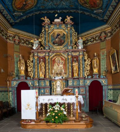 Zdjęcie nr 1: Kamienny ołtarz w jawiszowickim kościele uzyskał kształt prostopadłościanu. Otacza go drewniana obudowa podtrzymująca drewnianą mensę z portatylem. Jego przód zdobi drewniane antepedium ujęte profilowaną ramą z centralnym medalionem z monogramem Marii, otoczonym przez ornament roślinny. Ponad ołtarzem rozpościera się dwukondygnacyjna, architektoniczna nastawa ołtarzowa, flankowana przez dwie bramki. Struktura ołtarza dekorowana jest ciemnozieloną marmoryzacją. Pierwsza kondygnacja retabulum uzyskała trójosiowe założenie wsparte na dwustrefowym cokole, podzielonym płycinami z dekoracją akantową. Artykułują ją dwie pary kolumn o skręconych trzonach i kapitelach kompozytowych. Złocone w całości kolumny podtrzymują belkowanie o wysuniętym gzymsie, wyłamane na ich osi oraz przerwany trójkątny przyczółek. Pole środkowe nastawy wypełnia w całości nisza ujęta profilowaną, złoconą ramą w formie stojącego prostokąta zamkniętego trzema bokami z ośmioboku. W jej wnętrzu znajduje się obraz Matki Boskiej z Dzieciątkiem w typie Matki Boskiej Śnieżnej, zdobiony drewnianą złoconą i srebrzoną sukienką wotywną oraz koronami, a także dwie zasuwy z przedstawieniem św. Marcina, biskupa oraz bł. Salomei. Niszę środkową flankują znajdujące się pomiędzy kolumnami wąskie płyciny zwieńczone konchami. Na ich tle ustawiono figury św. Piotra i św. Pawła. Ponad nimi widoczne są na belkowaniu płaskorzeźbione i złocone girlandy kwiatowe oraz rozety. Na przerwanym przyczółku umieszczono dwie figury siedzących aniołów. Na mensie ołtarza centralnie ustawiono prostopadłościenne tabernakulum w drewnianej obudowie ujęte dwiema kolumienkami kompozytowymi o skręconych trzonach podtrzymujących belkowanie, do którego przymocowano świeczniki na sakramentki. Umieszczony na jego szczycie prosty tron eucharystyczny bez zaplecka flankują dwie figury uskrzydlonych aniołków. Drugą, jednoosiową kondygnację nastawy ujmuje także para złoconych kolumn o skręconych trzonach i kapitelach kompozytowych, ustawionych na cokołach, dźwigających imposty. Pomiędzy kolumnami nad polem z obrazem Wniebowzięcia Matki Boskiej widoczny jest wysunięty fragment gzymsu. Obraz ujęty został złoconą i profilowaną ramą w formie kwadratu o ściętych narożach. Po bokach obrazu na płycinie rozmieszczono złocone dekoracje akantowe. W zwieńczeniu na belkowaniu usytuowano dwa putta flankujące rzeźbę apokaliptycznego baranka leżącego na księdze. Obie kondygnacje ujmują uszaki z akantu suchego. Po bokach nastawy usytuowano dwie bramki flankowane pilastrami. Ich zwężające się ku dołowi trzony podtrzymują nadwieszony łuk półkolisty. Całość zamyka gzyms z centralnie usytuowanym cokołem, na którym stoją figury św. Stanisława i św. Wojciecha. Po ich bokach figur przy ścianach ustawiono rzeźbione i złocone wazony z kwiatami. 