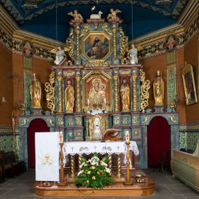 Zdjęcie nr 1: Kamienny ołtarz w jawiszowickim kościele uzyskał kształt prostopadłościanu. Otacza go drewniana obudowa podtrzymująca drewnianą mensę z portatylem. Jego przód zdobi drewniane antepedium ujęte profilowaną ramą z centralnym medalionem z monogramem Marii, otoczonym przez ornament roślinny. Ponad ołtarzem rozpościera się dwukondygnacyjna, architektoniczna nastawa ołtarzowa, flankowana przez dwie bramki. Struktura ołtarza dekorowana jest ciemnozieloną marmoryzacją. Pierwsza kondygnacja retabulum uzyskała trójosiowe założenie wsparte na dwustrefowym cokole, podzielonym płycinami z dekoracją akantową. Artykułują ją dwie pary kolumn o skręconych trzonach i kapitelach kompozytowych. Złocone w całości kolumny podtrzymują belkowanie o wysuniętym gzymsie, wyłamane na ich osi oraz przerwany trójkątny przyczółek. Pole środkowe nastawy wypełnia w całości nisza ujęta profilowaną, złoconą ramą w formie stojącego prostokąta zamkniętego trzema bokami z ośmioboku. W jej wnętrzu znajduje się obraz Matki Boskiej z Dzieciątkiem w typie Matki Boskiej Śnieżnej, zdobiony drewnianą złoconą i srebrzoną sukienką wotywną oraz koronami, a także dwie zasuwy z przedstawieniem św. Marcina, biskupa oraz bł. Salomei. Niszę środkową flankują znajdujące się pomiędzy kolumnami wąskie płyciny zwieńczone konchami. Na ich tle ustawiono figury św. Piotra i św. Pawła. Ponad nimi widoczne są na belkowaniu płaskorzeźbione i złocone girlandy kwiatowe oraz rozety. Na przerwanym przyczółku umieszczono dwie figury siedzących aniołów. Na mensie ołtarza centralnie ustawiono prostopadłościenne tabernakulum w drewnianej obudowie ujęte dwiema kolumienkami kompozytowymi o skręconych trzonach podtrzymujących belkowanie, do którego przymocowano świeczniki na sakramentki. Umieszczony na jego szczycie prosty tron eucharystyczny bez zaplecka flankują dwie figury uskrzydlonych aniołków. Drugą, jednoosiową kondygnację nastawy ujmuje także para złoconych kolumn o skręconych trzonach i kapitelach kompozytowych, ustawionych na cokołach, dźwigających imposty. Pomiędzy kolumnami nad polem z obrazem Wniebowzięcia Matki Boskiej widoczny jest wysunięty fragment gzymsu. Obraz ujęty został złoconą i profilowaną ramą w formie kwadratu o ściętych narożach. Po bokach obrazu na płycinie rozmieszczono złocone dekoracje akantowe. W zwieńczeniu na belkowaniu usytuowano dwa putta flankujące rzeźbę apokaliptycznego baranka leżącego na księdze. Obie kondygnacje ujmują uszaki z akantu suchego. Po bokach nastawy usytuowano dwie bramki flankowane pilastrami. Ich zwężające się ku dołowi trzony podtrzymują nadwieszony łuk półkolisty. Całość zamyka gzyms z centralnie usytuowanym cokołem, na którym stoją figury św. Stanisława i św. Wojciecha. Po ich bokach figur przy ścianach ustawiono rzeźbione i złocone wazony z kwiatami. 