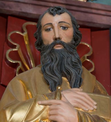 Zdjęcie nr 7: Pełnoplastyczna rzeźba św. Pawła Apostoła została ustawiona na zielonej podstawie na planie kwadratu o ściętych narożach. Święty został ukazany frontalnie w całej postaci. Apostoł ma nieznacznie pochyloną głowę do przodu i ręce skrzyżowane na piersi. Prawą podtrzymuję księgę wspartą o lewe przedramię, trzymając jednocześnie w lewej miecz, skierowany ostrzem ku ziemi. Święty Paweł ma pociągłą twarz okoloną czarnymi włosami i rozdwojoną, długą brodę. Jego spojrzenie skierowane jest w dół. Ma prosty nos, wyraziste oczy i usta. Ubrany jest w przylegającą do ciała tunikę oraz płaszcz narzucony na lewe ramię i podtrzymywany z przodu. Spod tuniki widoczne są bose stopy. W partiach ciała widoczna jest polichromia naturalistyczna. Natomiast szaty i atrybuty są złocone. 